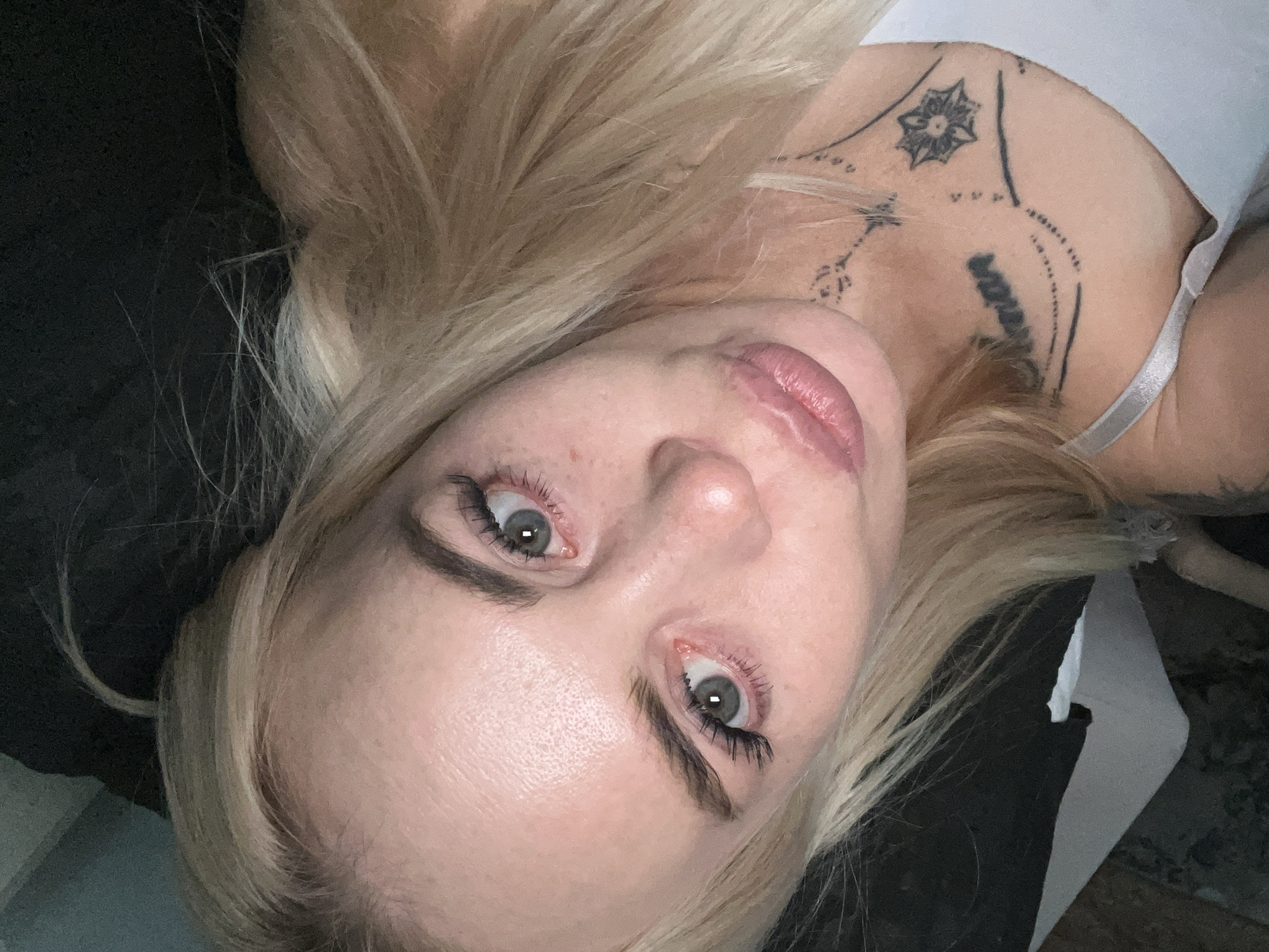 This is a close-up, upside-down, eye-level portrait of a young, fair-skinned woman with blonde hair. She has light gray-blue eyes with dramatic false eyelashes, and her eyebrows are dark and well-defined. Her lips are full and appear to be naturally pink. She is wearing light makeup, with some subtle blush on her cheeks.
On her right shoulder and collarbone area, she has several black tattoos. One prominent tattoo is a star-like, geometric design on her upper collarbone. Below that, there is some text that appears to be in black ink. A light-colored bra strap is visible on her right side.
The lighting suggests an indoor setting, possibly with artificial light, as there are no strong shadows indicative of natural sunlight. The background is dark and out of focus, with some blurry textures that could be fabric or furniture. The woman's expression is neutral, with her gaze directed towards the camera. The overall mood of the image is intimate and personal, focusing on the subject's face and upper body. The time of day and weather are not discernible. The location is unknown.
