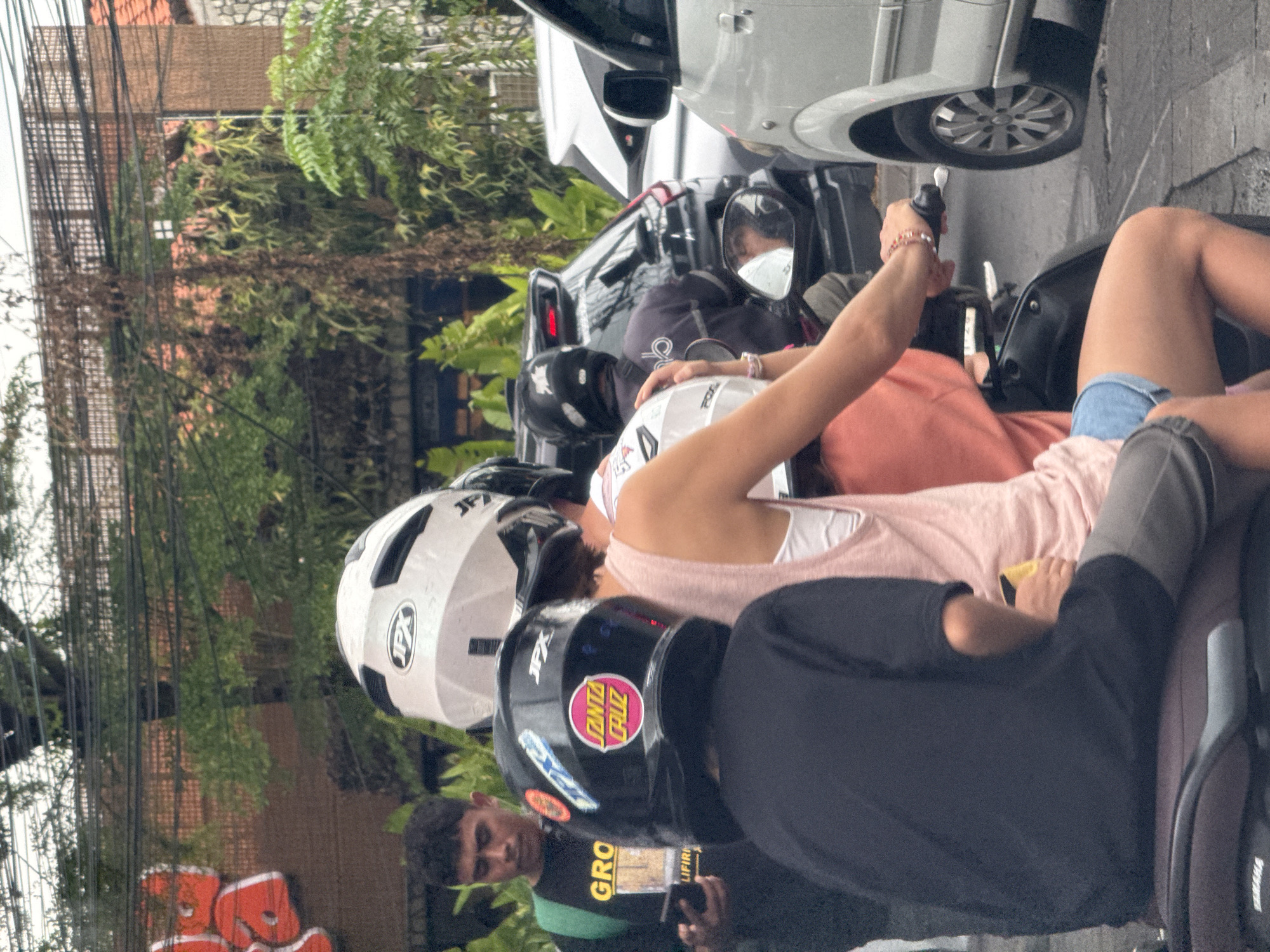 This vertical image depicts a street scene in North Kuta, Indonesia, with several people on motorcycles or scooters alongside a parked car. In the foreground, a person in a black helmet adorned with a "SANTA CRUZ" sticker and a black shirt is visible. Next to them, another individual wears a light pink top and denim shorts, holding onto a handlebar. Behind them, a person with a white helmet featuring "JPX" branding is seen, also in a pink top. Another rider wears a black helmet further back.
A silver car is parked on the right side of the frame, with a person wearing a white face mask visible in its side mirror. In the lower left, a young man wearing a green and black shirt with "GRO" and partially "LIFIRH" text, looks towards the camera while holding a phone.
The background is rich with dense green foliage and trees, interspersed with numerous dark power lines or cables. Buildings with orange-tiled roofs are partially visible through the greenery. The scene appears to be set during the daytime under diffused lighting, suggesting a moment of pause for the vehicles and individuals, characteristic of urban or semi-urban areas in tropical regions.