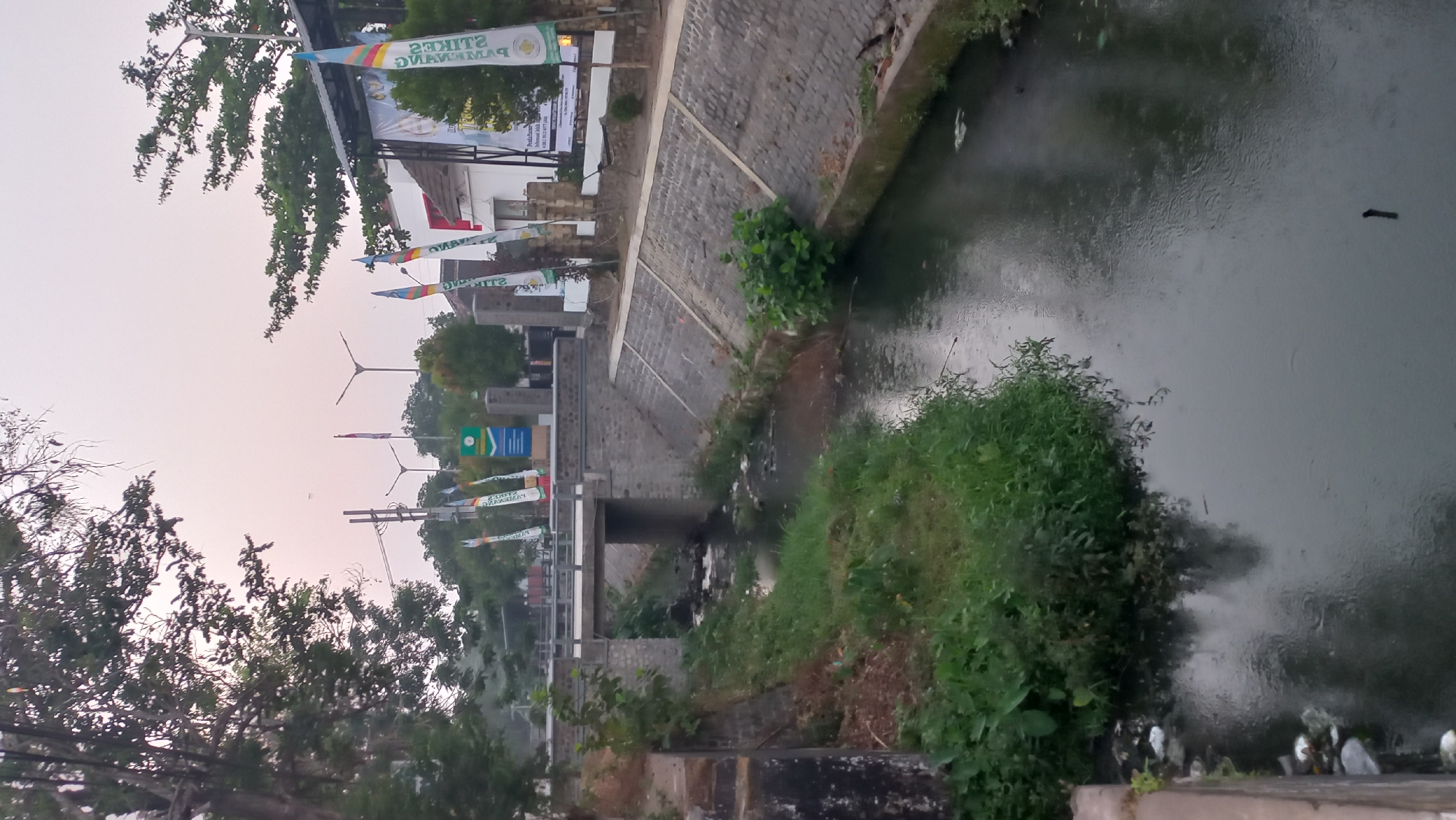 The image depicts a canal in the city of Pare, Indonesia. The canal's sides are reinforced with stone walls, and lush green vegetation grows along its edges. A bridge crosses the canal in the mid-ground, supported by stone pillars. In the background, buildings and streetlights are visible. Flags, some bearing text and graphics, are planted along the canal bank, suggesting a public event or decoration. The water in the canal appears murky and reflects the overcast sky. Litter is present along the canal's edge on the left side of the image. The overall scene suggests a functional urban waterway with elements of public display and a degree of environmental neglect.