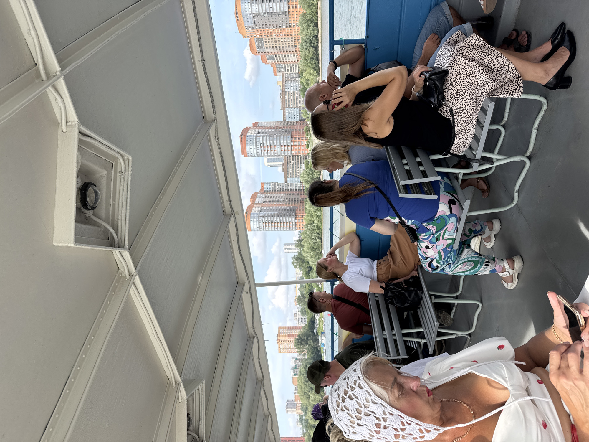 On a bright, sunny day, a group of people are seated on a ferry boat. The ferry is moving on a river with a cityscape in the background. The city is characterized by tall, modern buildings with orange and gray facades, nestled amongst lush green trees. The sky above is a clear blue with scattered white clouds.
The people on the ferry are casually dressed, enjoying a leisurely boat ride. On the left, a woman with blonde hair and a white crocheted headscarf is looking at her phone. She is wearing a white dress with red strawberry prints. To her right, a man in a green cap and a red shirt is seated, facing away from the camera. Next to him, a woman with blonde hair in a white t-shirt and a brown skirt is looking out at the water. Further down the row, a woman in a blue top is seated next to a woman with blonde hair in a grey top, and a man in a black t-shirt. A woman in a black tank top and a patterned skirt is talking on her phone, seated next to a man in a blue shirt and denim shorts.
The interior of the ferry is visible, with a white, ribbed ceiling and blue seats. The floor appears to be made of a gray material. The ferry is equipped with benches for passengers to sit on. The overall atmosphere is relaxed and summery.