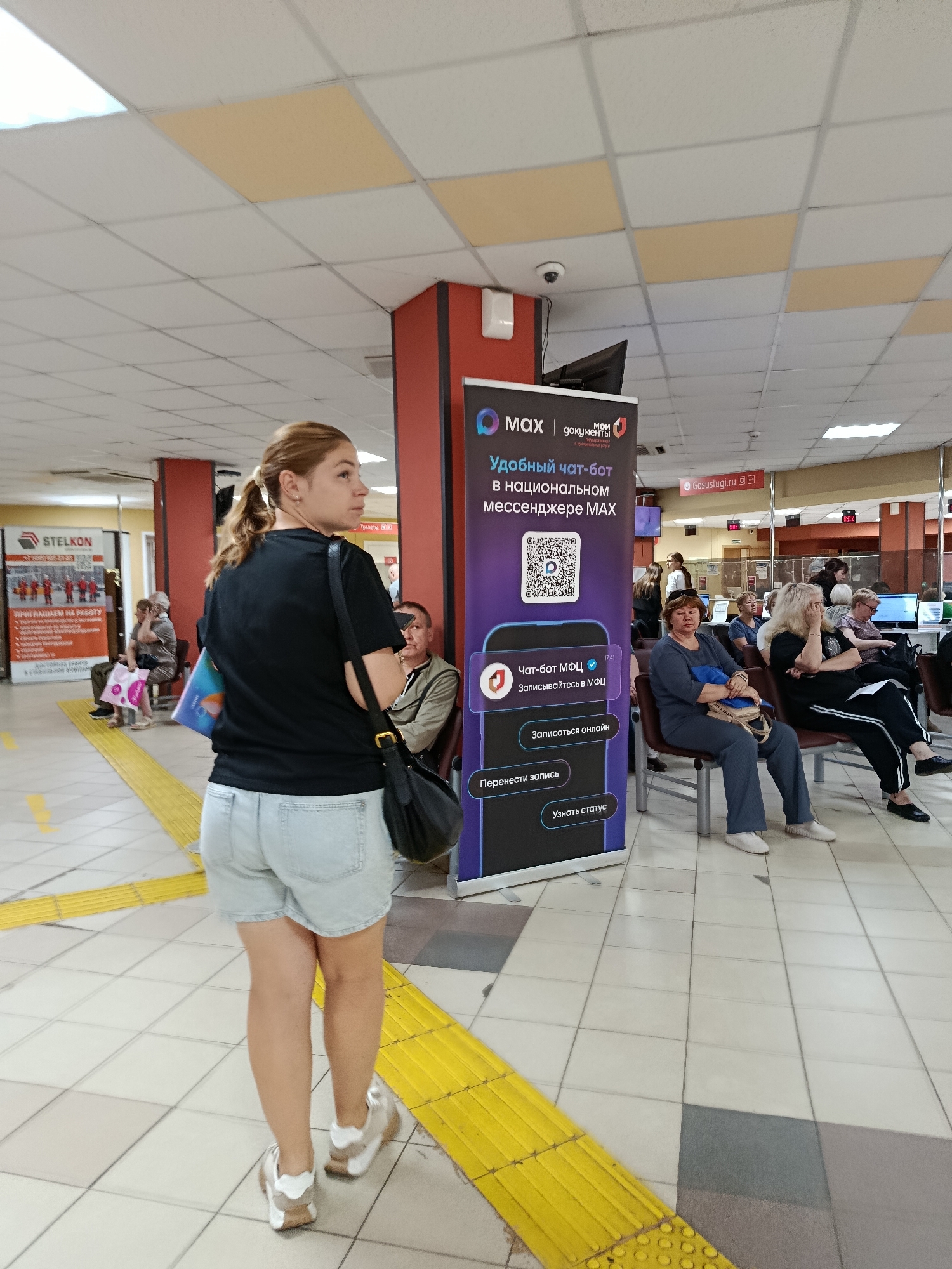 The image shows the interior of a public service center, likely a multifunctional center (MFC) in Orekhovo-Zuyevo, Russia. In the foreground, a young woman with blonde hair pulled back in a ponytail is seen from the back, looking over her right shoulder. She is wearing a black t-shirt and light blue denim shorts, with white and tan sneakers. She is carrying a black shoulder bag and holding a blue and white object.
Several other people are seated in a waiting area. On the right, a group of women and men are sitting on chairs. One woman in the front is wearing a dark blue tracksuit. Another woman to her right has blonde hair and is wearing dark pants with white stripes. A man in a light brown jacket is seated further back.
A large banner is prominently displayed on the left side of the image. The banner features the logo "MAX" and "МОИ ДОКУМЕНТЫ" (My Documents) and promotes a chatbot service. The banner has a QR code and text in Russian, including "Удобный чат-бот в национальном мессенджере MAX" (Convenient chatbot in the national messenger MAX) and options to "Записаться онлайн" (Sign up online), "Перенести запись" (Reschedule appointment), and "Узнать статус" (Check status).
Another banner on the far left has "STELKON" branding and images of workers. The environment is a brightly lit indoor space with tiled flooring and a suspended ceiling. Yellow tactile paving is visible on the floor, indicating accessibility features. The overall impression is a busy, public administrative building.