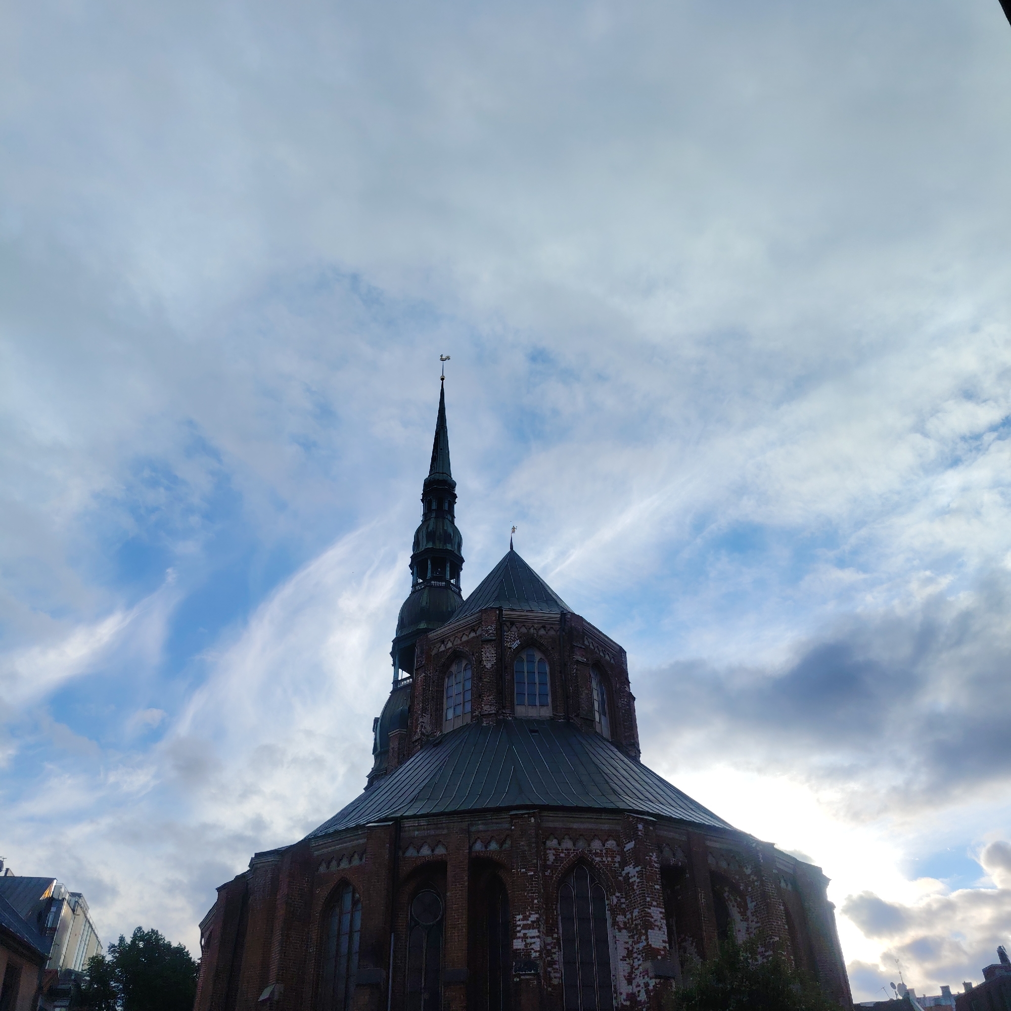 The image shows a low-angle view of the St. Peter's Church in Riga, Latvia. The church is constructed of red brick and features a tall, slender spire topped with a weather vane. The spire is dark green and has a series of progressively narrower sections as it ascends. Several arched windows are visible on the church's octagonal base. The sky above is filled with dramatic, wispy clouds, with patches of blue sky showing through. The lighting suggests it is either early morning or late afternoon. In the foreground, to the left of the church, part of another building with a darker roof is visible, along with some dark green foliage. The image is sharp and well-exposed.