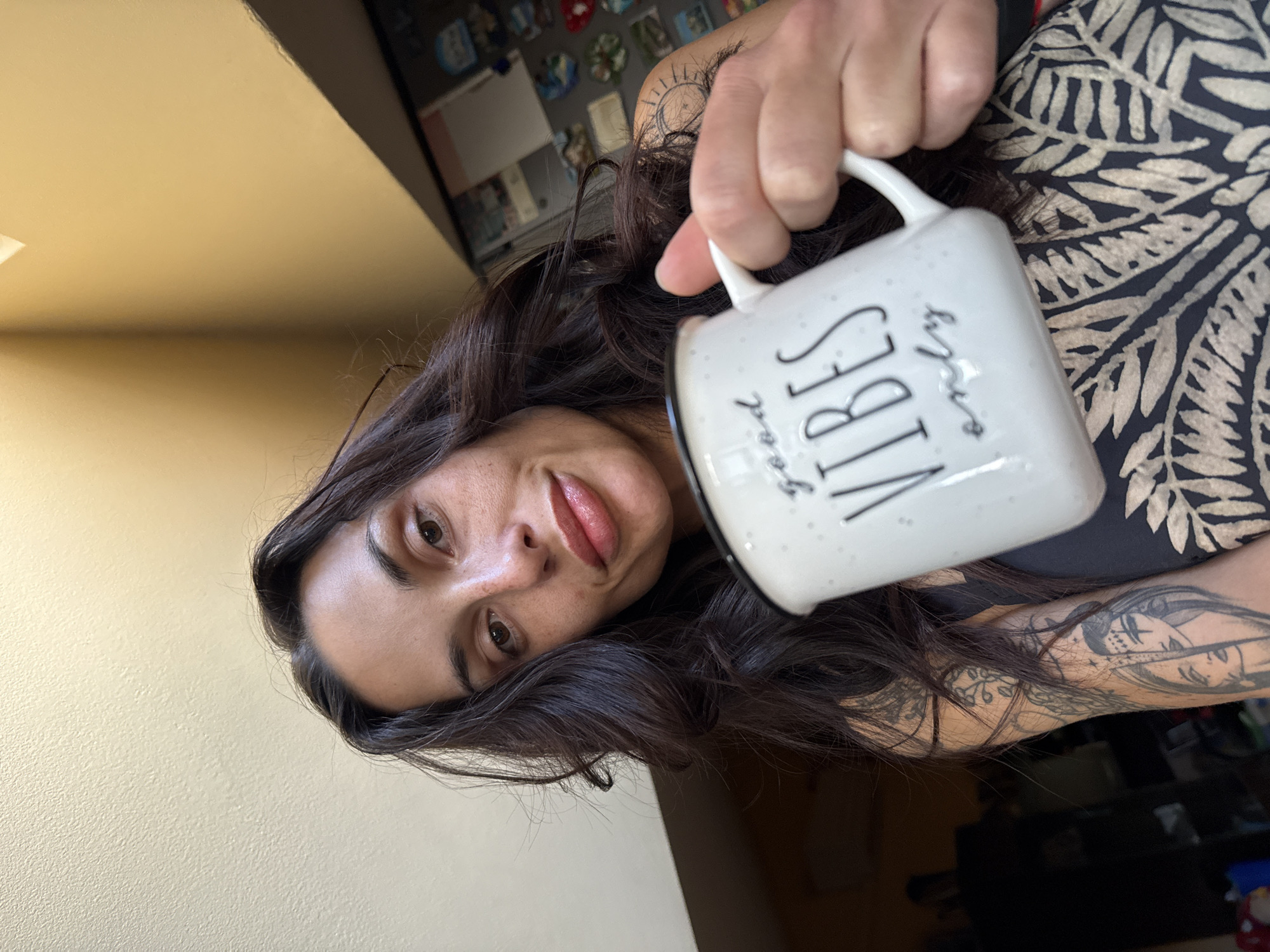 A woman is holding a white mug with black lettering that reads "Good Vibes Only". The woman has long, dark hair and is wearing a black and white patterned shirt. She has tattoos on her arm. The background includes a wall and a refrigerator with magnets. The location is likely indoors, but there is no information to identify the specific location as Pleven, Bulgaria.