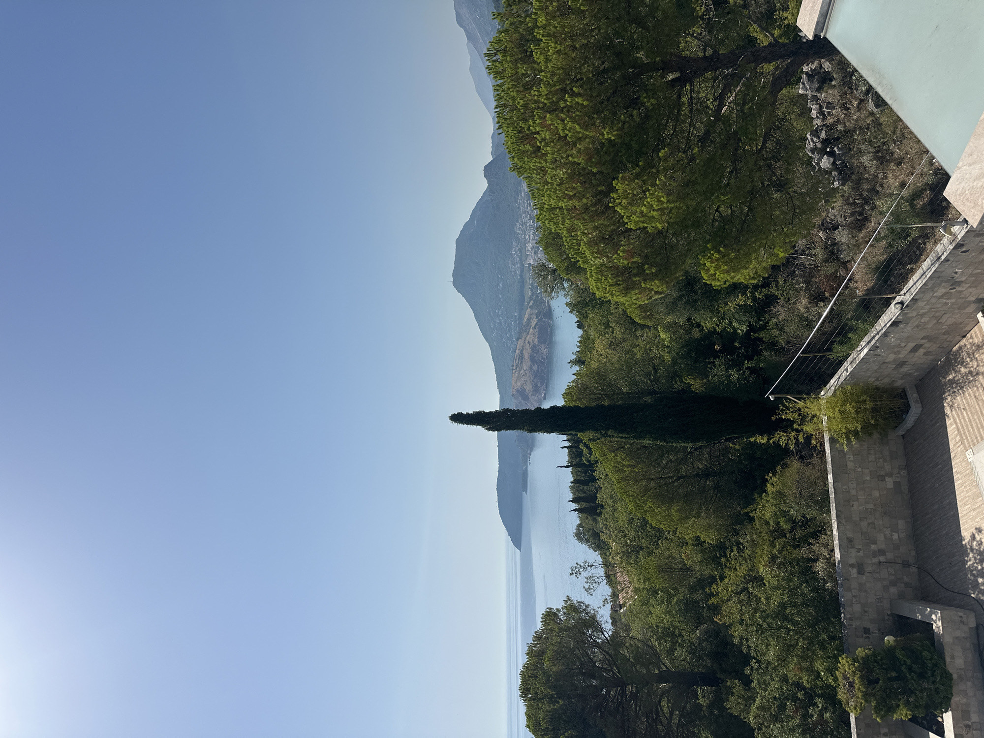 The image depicts a scenic view of the coastline near Brca, Montenegro. In the foreground, a patio area constructed with light-colored stone and wood-like tiles is visible. A metal railing separates the patio from a vegetated slope. Lush green trees and a tall, slender cypress tree dominate the mid-ground, with a variety of deciduous trees and shrubs creating a dense forest canopy. Beyond the trees, the clear blue Adriatic Sea stretches towards the horizon, meeting a hazy sky. A mountainous peninsula juts out into the sea on the left side of the frame, and in the distance, a larger mountain range is visible. A small town with buildings clustered along the coast can be seen nestled at the base of the mountains on the right. The overall atmosphere is tranquil and natural, with the sun casting shadows and highlighting the vibrant greenery. There are no people or animals visible in the image.