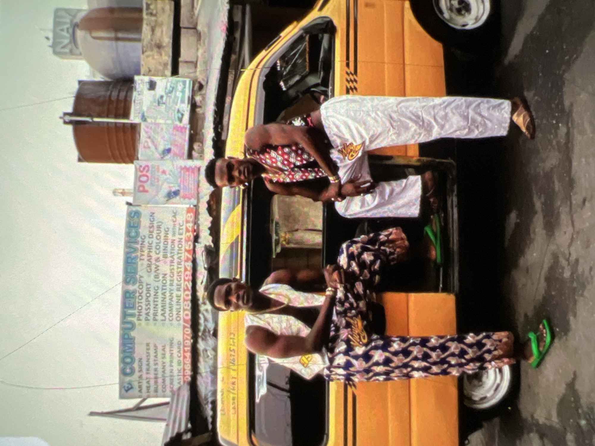The image depicts two young Black men posed in front of a yellow commercial van, likely a danfo, on a street in Lagos, Nigeria. The van has a sign above it reading "COMPUTER SERVICES" with a list of services including "PHOTCOPY, TYPING, GRAPHIC DESIGN, PRINTING (B/W & COLOUR), LAMINATION, BINDING, COMPANY REGISTRATION ETC." and phone numbers. The men are dressed in vibrant, patterned clothing. The man on the left wears a white vest over a patterned shirt and brightly colored patterned trousers. He is seated on the open door of the van. The man on the right wears a patterned vest over a bare chest and loose-fitting white trousers. He is leaning against the van. Both men are wearing sandals. In the background, there are other signs, water tanks, and the general urban environment of Lagos. The street appears to be wet, suggesting recent rain.