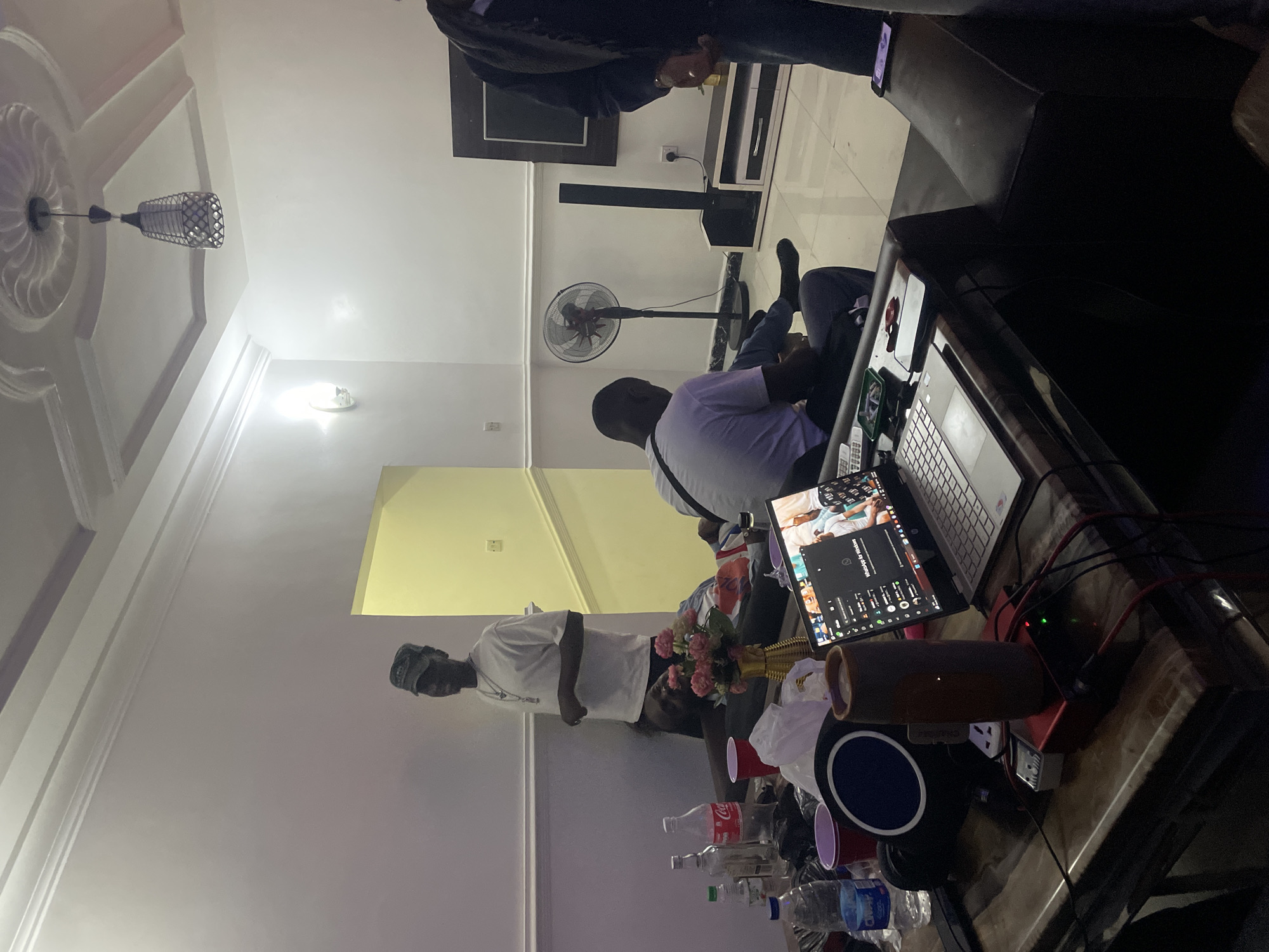 The image depicts an indoor gathering in a residential setting, likely in Galadimawa, Nigeria. In the foreground, a large marble-topped table holds various items including a laptop displaying WhatsApp for Windows, a cylindrical speaker, red cups, bottles of water and Coca-Cola, and other electronics. Two individuals are seated on a dark leather couch in the right foreground, with one partially visible from the side. In the mid-ground, a man wearing a white t-shirt and a cap stands near a doorway leading to a yellow-painted hallway. Another man in a grey t-shirt is seated on the floor facing away from the camera, with his legs extended. A standing fan is visible in the background near a television and entertainment unit. The room appears to be a living area, with white walls, a decorative ceiling with a light fixture, and tiled flooring. The overall atmosphere suggests a casual social event.
