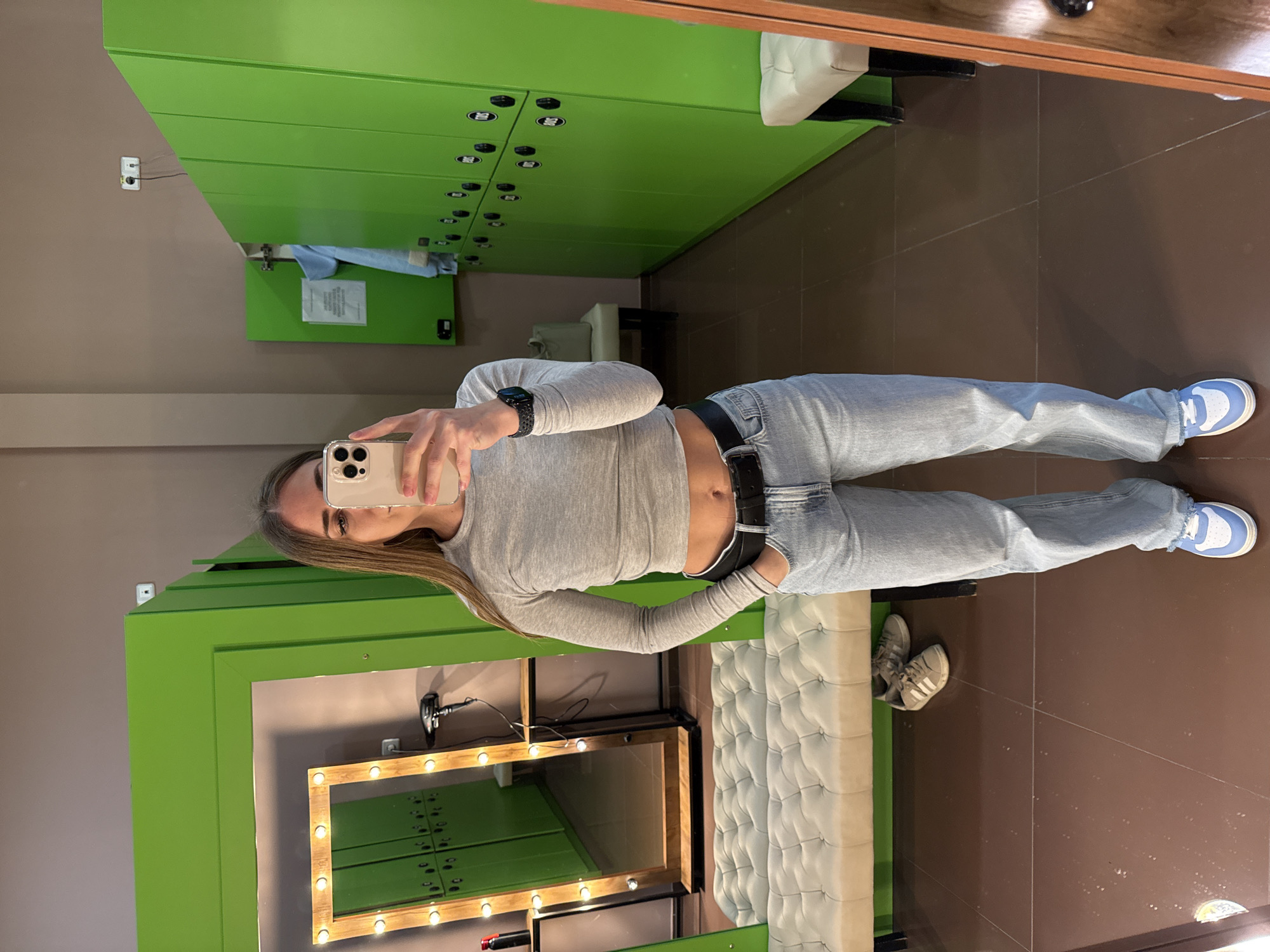 A young woman is captured in a mirror selfie within what appears to be a changing room or studio setting. She has long, light brown hair and is looking towards the phone she holds, partially obscuring her face. She is wearing a light gray long-sleeved crop top, light blue jeans, a black belt, and light blue and white sneakers. A black smartwatch is visible on her left wrist.
The background features bright green locker units lining the walls, some with circular black handles or locks. On a lower locker door, a white piece of paper with unreadable text is attached. A light brown wall with an electrical outlet and wires is also visible. The floor is covered with dark brown tiles.
To the right, a large vanity mirror with a light-colored wooden frame and multiple round light bulbs illuminates the scene. Reflected in this mirror, more green lockers can be seen. Below the mirror, a light-colored, tufted bench or seating area extends along the wall. Near the bench, on the tiled floor, another pair of gray sneakers (possibly Adidas) are visible. A black hairdryer or similar device rests on the surface next to the lighted mirror. The overall lighting suggests an indoor environment.