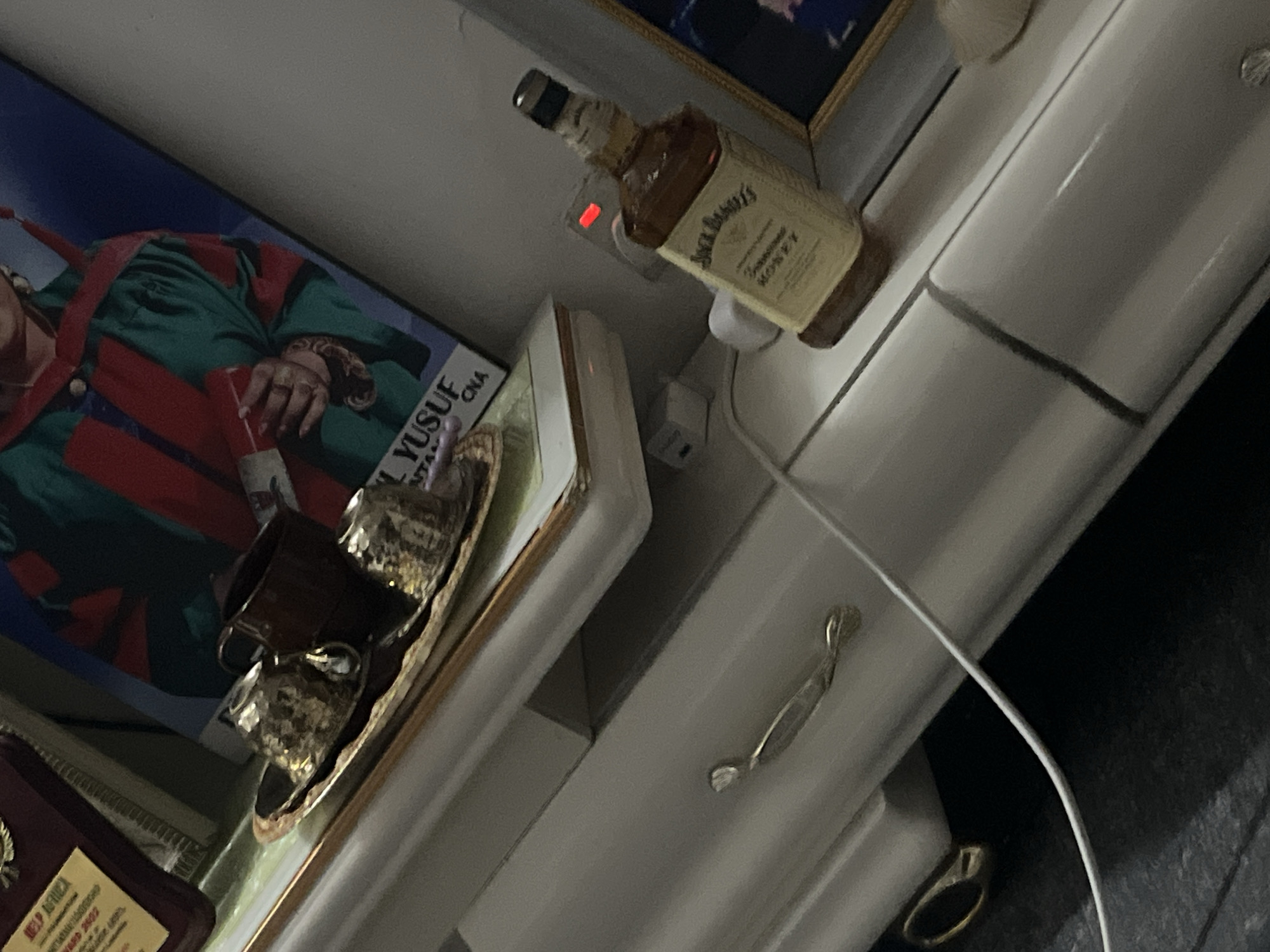 The image depicts a cluttered scene. A bottle of Jack Daniel's Tennessee Honey whiskey is plugged into a wall outlet. Below, there is a framed picture of a person identified as "Yusuf." A decorative tray with two small cups sits near a plaque or award, part of which reads "HELP AFRICA." The environment appears to be a domestic interior, likely in Lokoja, Nigeria, though no specific landmarks are visible.