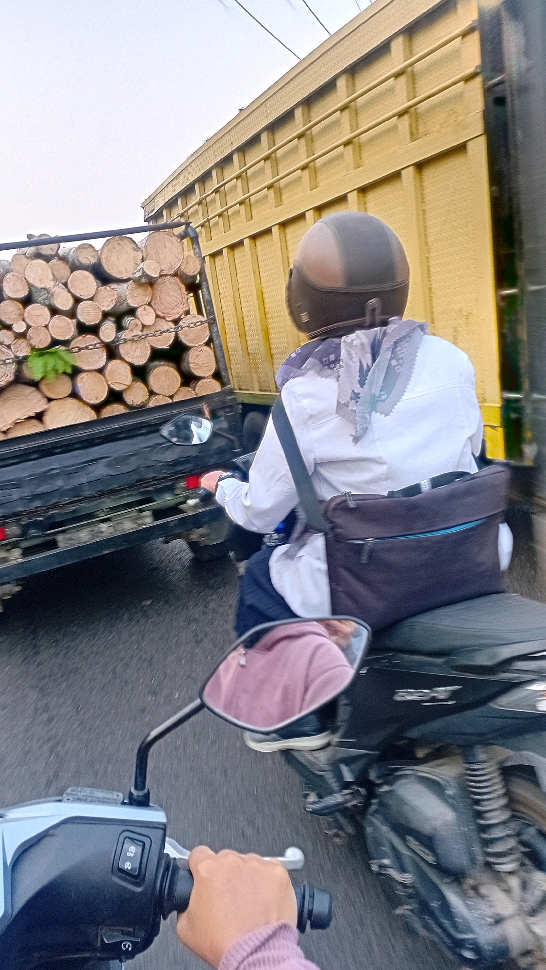 The image captures a street scene in Jambi, Indonesia, from the perspective of someone riding a motorcycle. In the foreground, a hand is visible on the handlebars of a motorcycle, with a pink sleeve on the arm. A rear-view mirror reflects the rider of the motorcycle in front, who is wearing a pink hooded sweatshirt. The primary subject is a person riding a motorcycle, wearing a helmet and a light-colored jacket with a patterned scarf. This person has a dark bag slung across their body. They are riding alongside a flatbed truck heavily loaded with logs. The truck is yellow and appears to be in motion, as does the motorcycle carrying the viewer. The road surface is visible and appears to be asphalt. The overall scene suggests a moment of transit on a road likely within the city of Jambi.