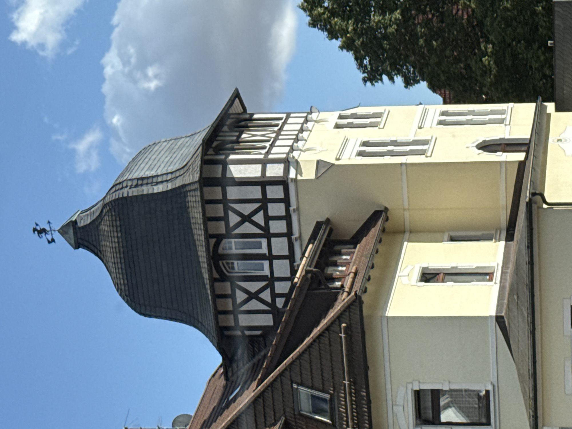 The image shows a close-up, eye-level view of a historic building in Bad Sachsa, Germany. The building features a prominent tower with a dark, mansard-style roof topped by a weather vane shaped like a horseman. The tower's upper section is adorned with traditional half-timbering in white and dark brown. The lower sections of the building are painted a pale yellow and have white decorative trim around the windows. Several windows are visible, including a narrow arched window on the tower and rectangular windows with white frames on the lower parts of the building. To the left of the tower, a steeply pitched roof covered in dark shingles is visible, with a smaller dormer window. A large, dark green tree partially obscures the right side of the building. The sky is blue with scattered white clouds. The lighting suggests it is daytime.