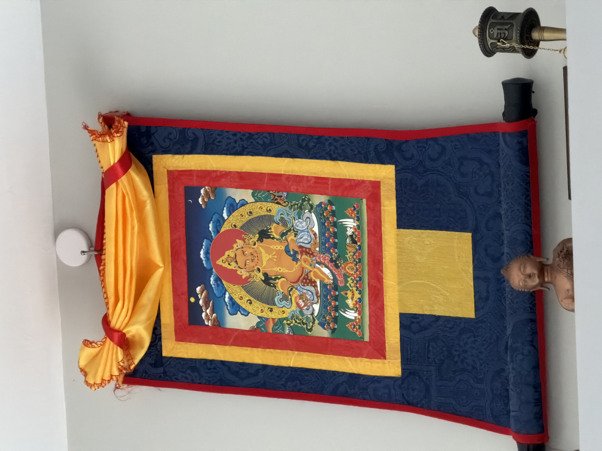 This media file depicts a decorative Buddhist scroll hanging on a wall, possibly in a home or a spiritual space in Florianópolis, Brazil. The scroll features a colorful illustration of a seated deity, likely a Buddhist figure, with intricate details and vibrant colors. The deity is adorned with jewelry and sits within a halo-like circular design, surrounded by symbolic imagery. The scroll itself is made of dark blue fabric with a subtle embossed pattern, bordered by bright red and yellow fabric trim. A section of bright yellow fabric is also incorporated into the design.
On the left side of the scroll, a piece of bright yellow fabric is draped and tied with a red ribbon, creating a decorative flourish. On the right side, a small, bronze-colored statue of a Buddha's head, with a serene expression, is partially visible, peeking out from behind a white pillar. At the top of the image, a Tibetan prayer wheel is mounted on the wall, suggesting a Buddhist cultural context. The overall setting appears to be indoors, with a clean, white wall as the backdrop. The lighting is even, suggesting it could be daytime with natural light, or a well-lit interior. There is no visible text in the image. The scroll and the prayer wheel suggest a spiritual or decorative display of Buddhist art and practice.