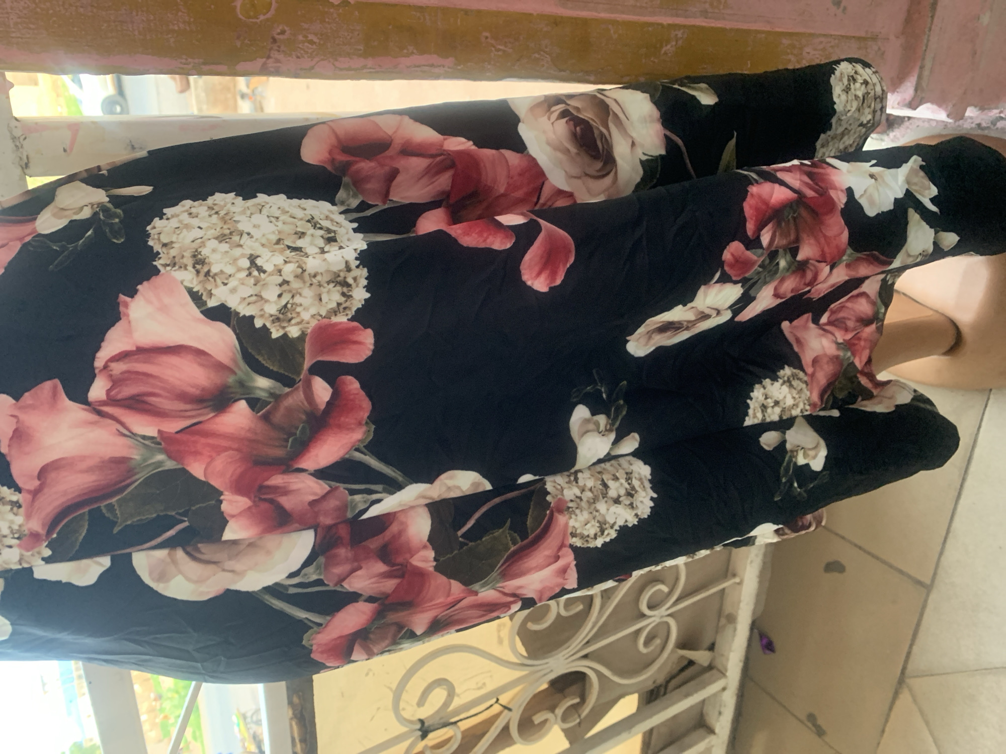 The image shows a close-up of a black dress with a floral pattern featuring large pink and white flowers. The dress appears to be made of a light, flowy material. The background is somewhat blurred but suggests an outdoor setting. To the left, there is a white metal railing or fence with decorative scrollwork. Behind the railing, some greenery and a street scene with vehicles are visible, indicating an urban environment. To the right, a textured, pale pink pillar or wall is present. The floor in the foreground appears to be tiled.
The scene is likely located in Jalingo, Nigeria, based on the provided context. The overall impression is of a fashion item displayed or worn outdoors, with a backdrop that suggests the everyday environment of the city. There are no people clearly visible in the image, only the dress and its surroundings. The lighting suggests it is daytime.