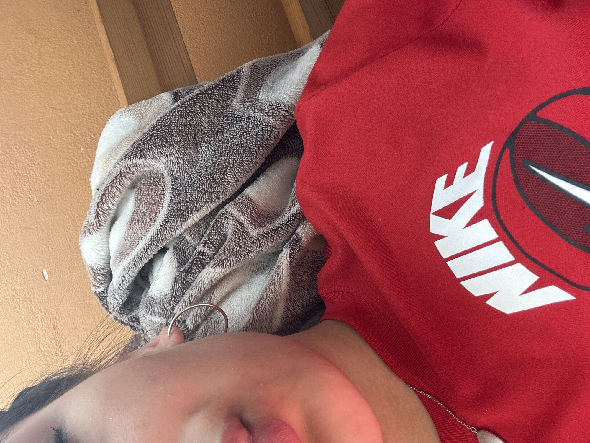 A close-up, indoor, eye-level shot captures the left side of a person's face and shoulder, along with a folded blanket and a portion of a red Nike t-shirt. The person's left eye, ear, and lips are visible, with a silver hoop earring in the ear. A thin silver chain with a heart-shaped pendant rests on the t-shirt. The blanket, draped over the person's shoulder, features a brown and white patterned design. The background consists of a textured orange wall and wooden planks. The scene appears to be located in a residential setting in Shumen, Bulgaria.