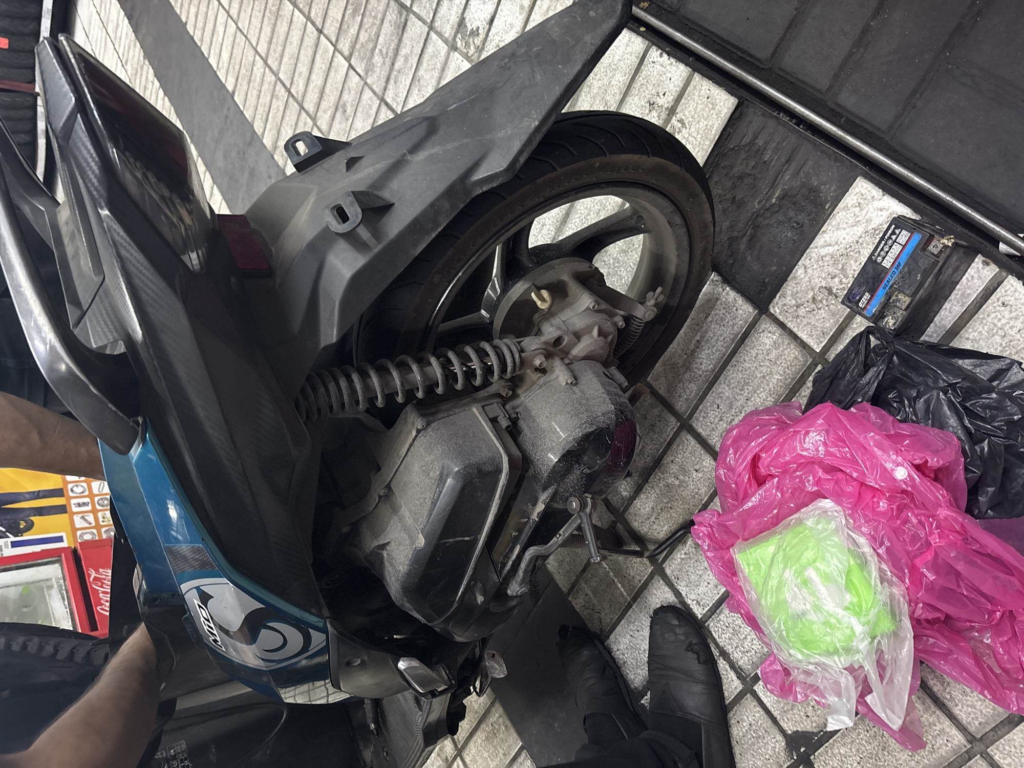 The image shows a close-up view of the rear section of a scooter, possibly in a repair shop, on a checkered tiled floor. The scooter is predominantly black with teal blue accents and a carbon fiber pattern on the rear fender and tail light housing. A person's legs and arms are visible near the front of the scooter, suggesting they are working on it.
Scattered on the floor are several plastic bags, one bright pink and another containing a green item. A used motorcycle battery with warning labels is also present near the bottom right of the frame. In the background, a red Coca-Cola branded cooler is visible, along with stacks of tires. The overall environment appears to be an urban commercial space, likely a tire shop or a motorcycle service center. While the specific location in North Jakarta, Indonesia cannot be definitively identified from the image alone, the presence of the Coca-Cola branding and the type of establishment are consistent with such a setting in that region.