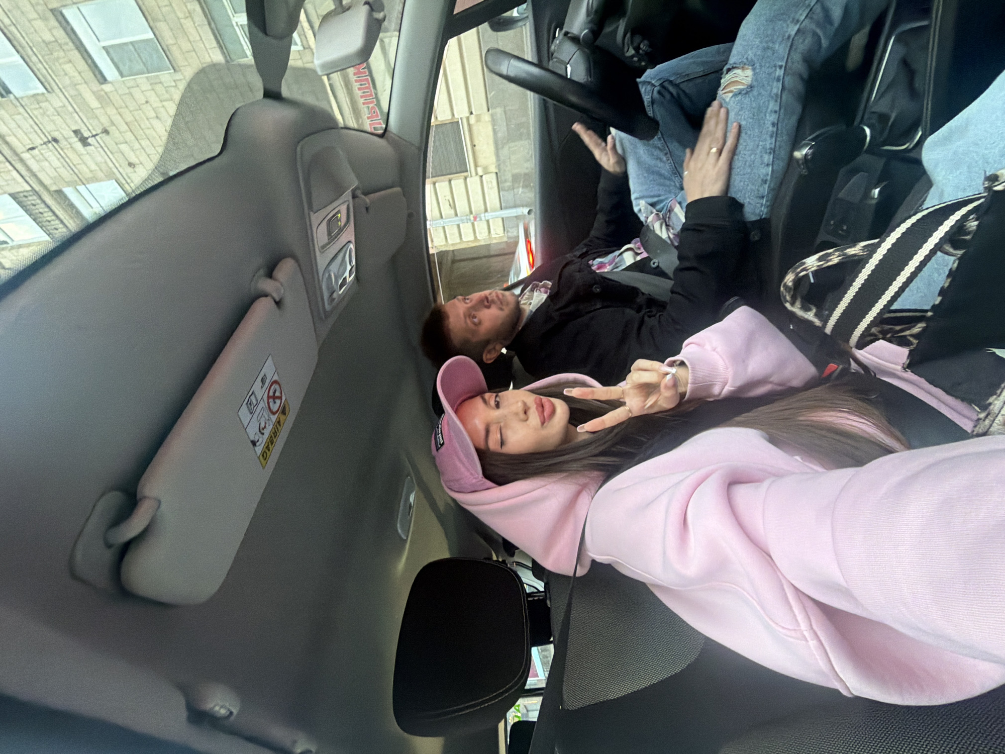 The image shows a couple inside a vehicle, likely a car, in Saint Petersburg, Russia, as discerned from the building visible through the windshield. The woman is in the passenger seat, wearing a pink hoodie and cap, and is making a peace sign with her fingers towards the camera. The man, presumably the driver, is wearing a black jacket over a patterned shirt. The car's interior is visible, including the dashboard, steering wheel, sun visor, and seats. A building exterior is visible through the car windows.