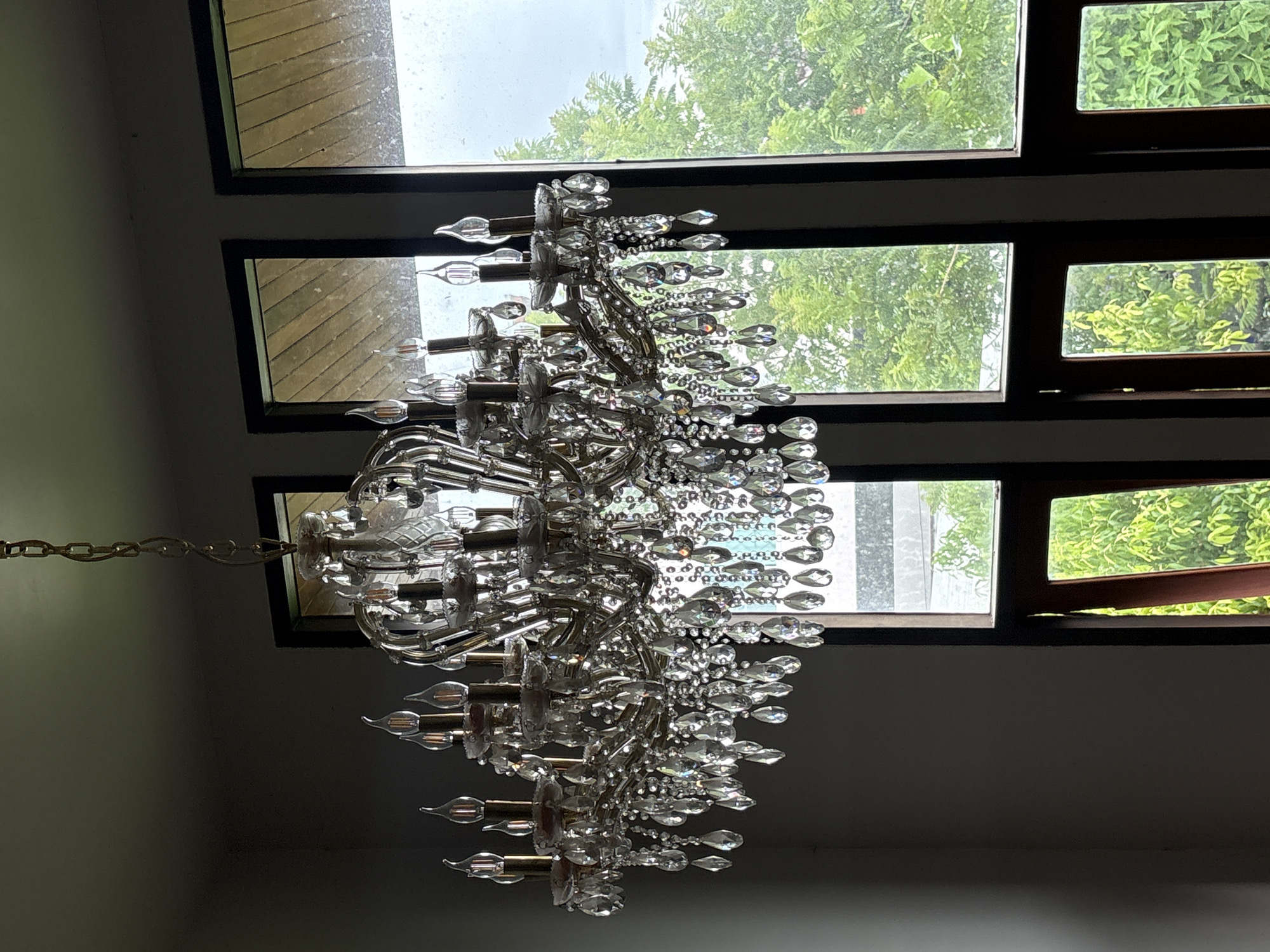 This media file shows a close-up view of a crystal chandelier hanging from a chain. The chandelier is ornate, with multiple tiers of crystal droplets and candle-shaped light fixtures. It is suspended against a backdrop of windows that look out onto lush green trees and a cloudy sky, suggesting an outdoor, possibly humid, environment. The lighting conditions are dim, indicating it might be daytime with overcast weather. The setting appears to be indoors, given the chandelier and ceiling structure. There are no people visible in the frame. The image is taken from a low angle, looking upwards at the chandelier. The location context provided is "บ้านกาญจน์กนก 23, Thailand".