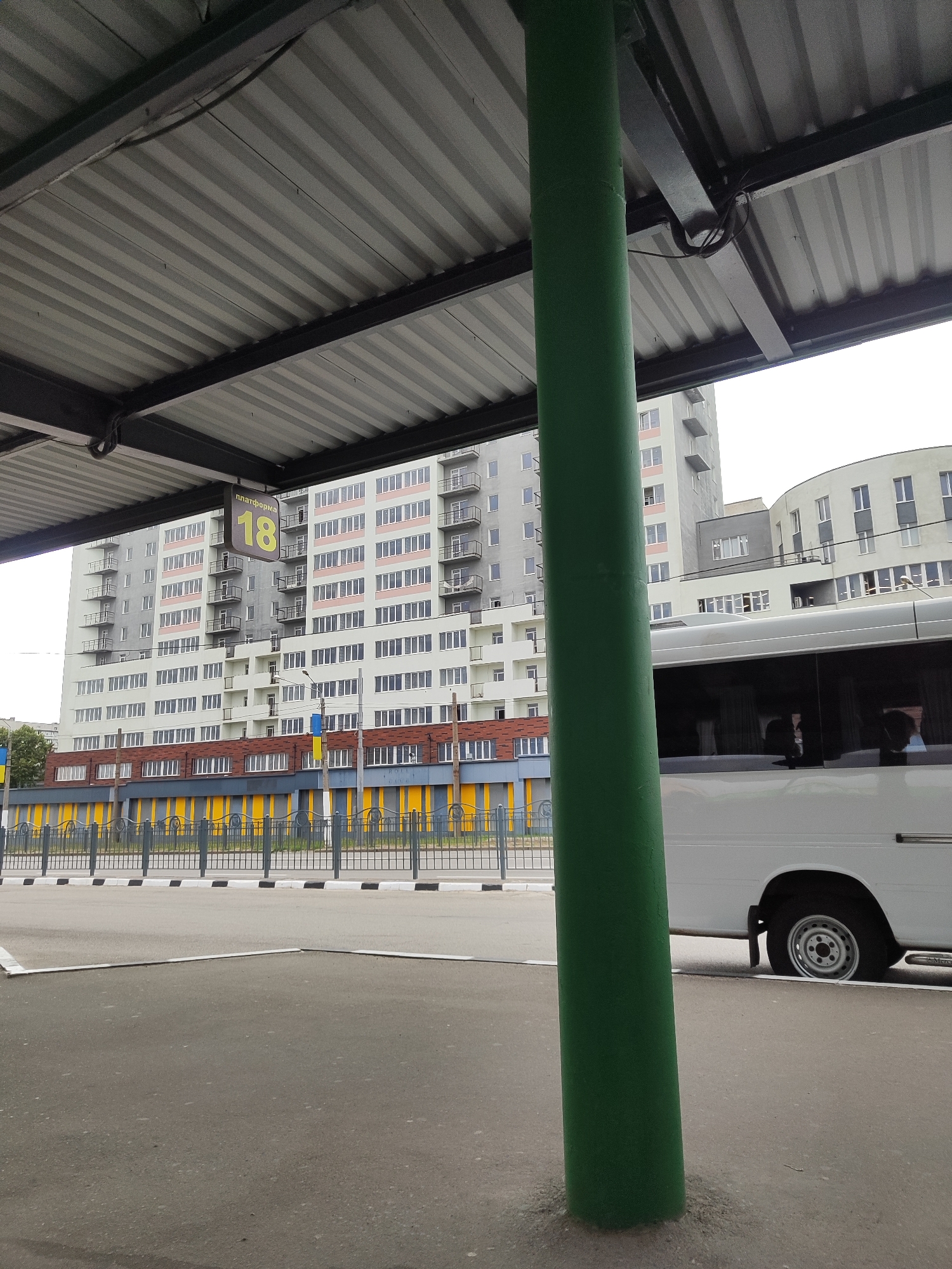 A bus stop shelter with a corrugated metal roof is visible, supported by a thick green pillar in the foreground. A sign indicating "ПЛАТФОРМА 18" (Platform 18) hangs from the shelter. In the background, a modern apartment building with multiple balconies dominates the scene. A white bus is parked at the bus stop, its windows reflecting the surroundings. A yellow and blue striped fence runs along the front of the apartment complex. The ground consists of asphalt pavement and a sidewalk. The sky is overcast.