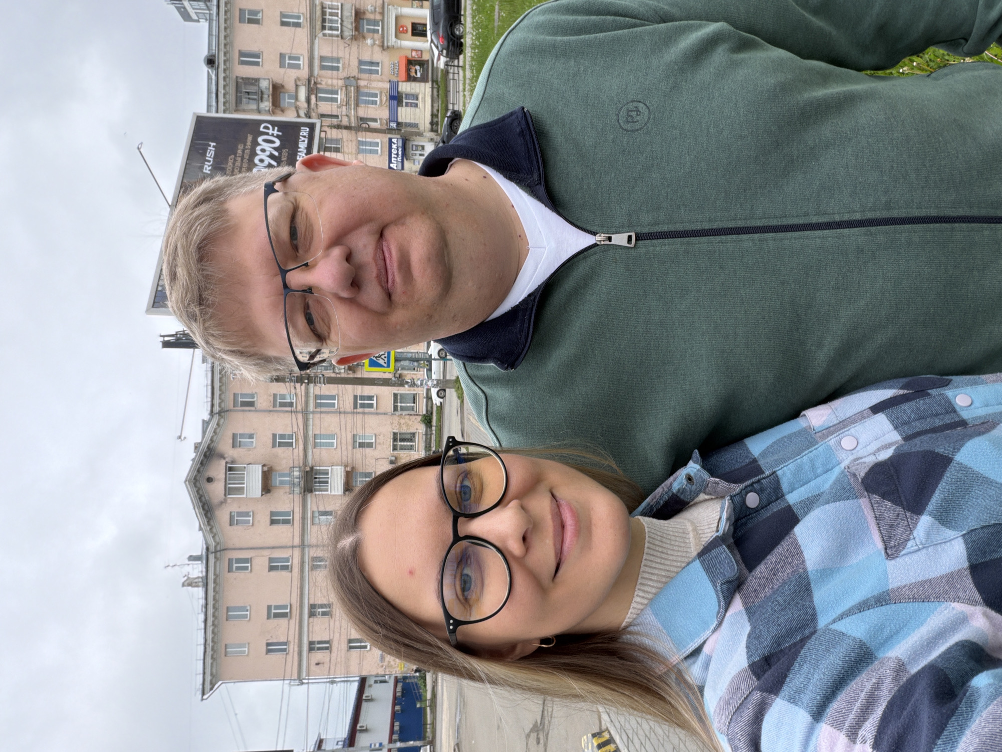 An outdoor portrait captured in daylight features a man and a woman posing together, likely a selfie, in an urban setting. The sky is overcast.
The man, positioned on the left, wears black-framed glasses and a dark green zip-up jacket over a white shirt. A small, circular logo is visible on his jacket's left chest. He has light-colored hair and a neutral expression.
The woman, on the right and slightly in front, also wears black-framed glasses. She has light brown hair and a gentle smile, dressed in a blue, gray, and white plaid button-up shirt over a light-colored ribbed top.
In the background, behind the subjects, are several multi-story buildings, consistent with a residential or mixed-use area. A large billboard stands prominently, displaying text including "RUSH," "990₽" (990 Russian Rubles), and other Russian words such as "САМЫЙ СМЕЛЫЙ" (The bravest), "ПОКУПАЙ ТЫ" (Buy you), "НАШ ТОВАР" (Our product), and "FAMILY.RU." Further in the background, a building has a sign that reads "АПТЕКА Плюс" (Pharmacy Plus). A road with visible markings, and patches of grass are also discernable. The image orientation is sideways. The scene depicts a casual moment in Izhevsk, Russia.