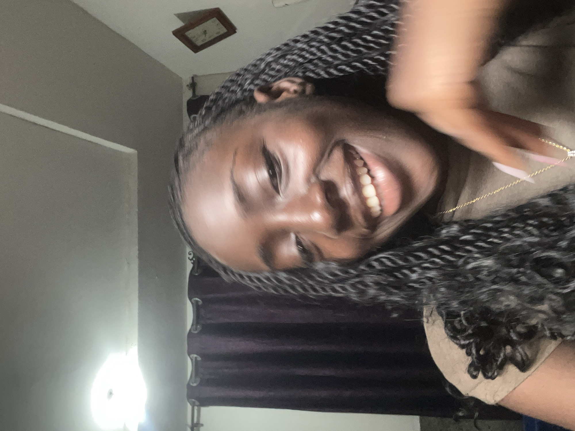A young woman with dark skin and long, black braided hair smiles broadly at the camera. She is wearing a tan-colored shirt and a gold chain with a pendant. Her left hand is visible in the foreground, slightly blurred, and appears to be touching her chest. The background consists of dark purple curtains and a wall with a framed picture. The lighting suggests an indoor setting, possibly a bedroom or living room. The environment appears to be a residential area within the city of Abuja, Nigeria.