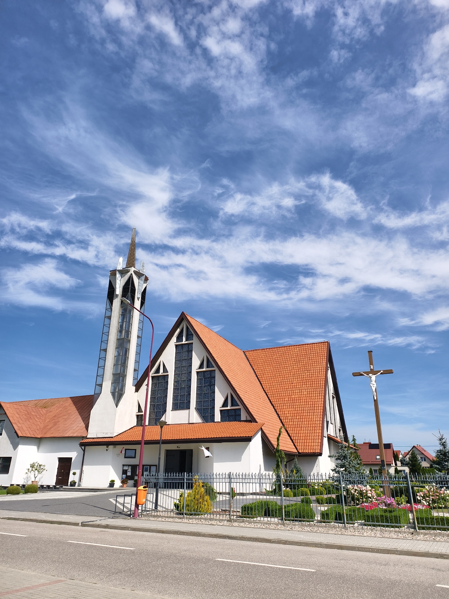 The image shows the Church of the Holy Spirit in Tczew, Poland, under a bright blue sky with wispy white clouds. The modern church features a prominent, tall, white bell tower with a pointed spire. The main church building has an orange tiled roof and large, arched windows with frosted glass panes. To the right of the church, a wooden crucifix with a white figure of Jesus Christ stands prominently. The church is surrounded by a neatly manicured garden with bushes, small trees, and colorful flowers, enclosed by a black metal fence. A paved road and sidewalk are visible in the foreground. The area appears to be a residential neighborhood, with houses visible behind the church.