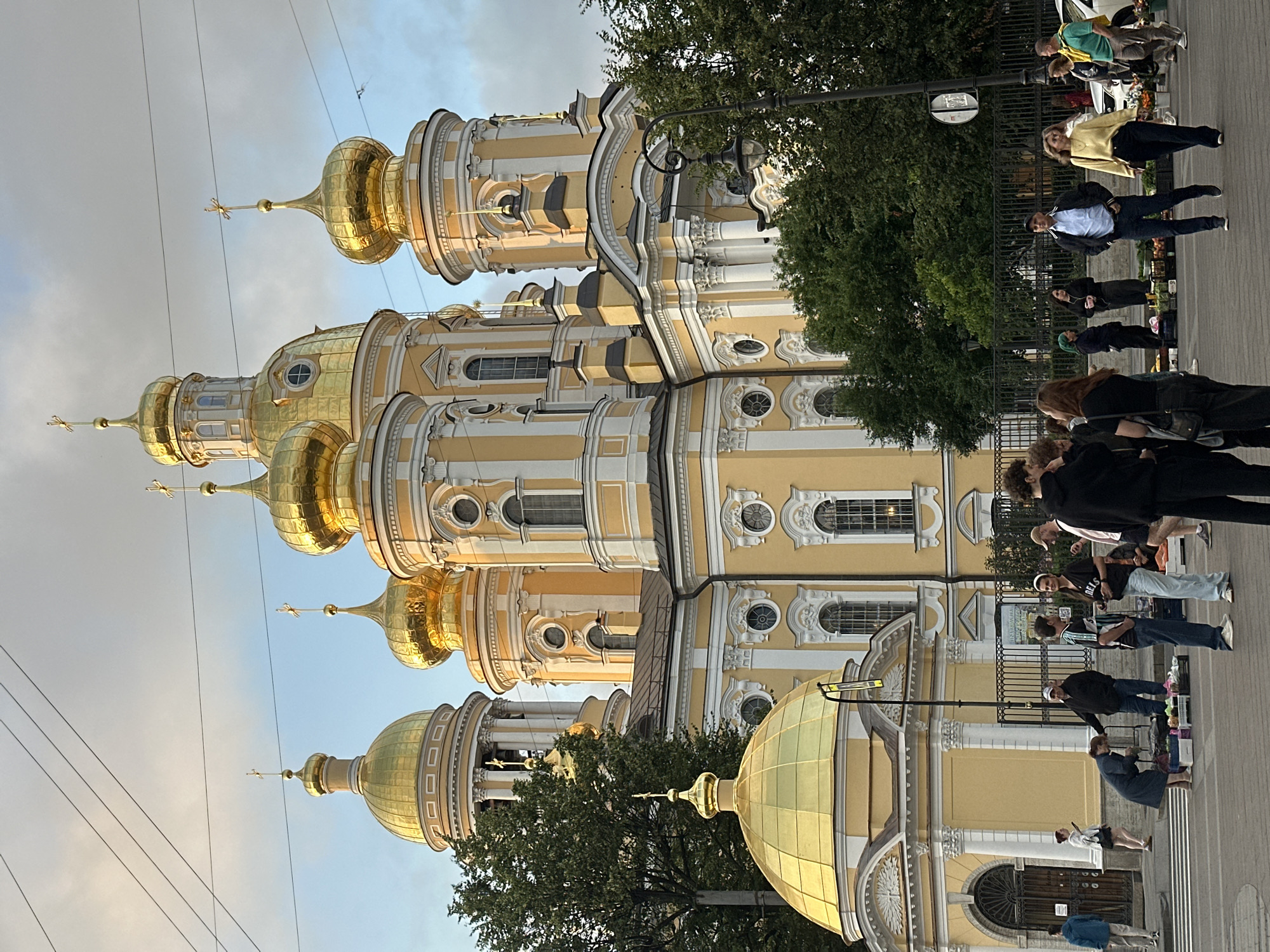 This is a daytime, eye-level, medium shot of the Smolny Cathedral in St. Petersburg, Russia. The cathedral, an example of Elizabethan Baroque architecture, features a prominent yellow facade with white decorative elements and multiple golden domes topped with crosses. In the foreground, a group of approximately ten people are gathered on a paved area, some interacting with each other and others appearing to browse merchandise laid out on crates. The people are dressed in casual clothing. Trees flank the cathedral and the immediate area. Overhead power lines are visible against a cloudy sky. The scene suggests an urban environment with a mix of historical architecture and contemporary street life.