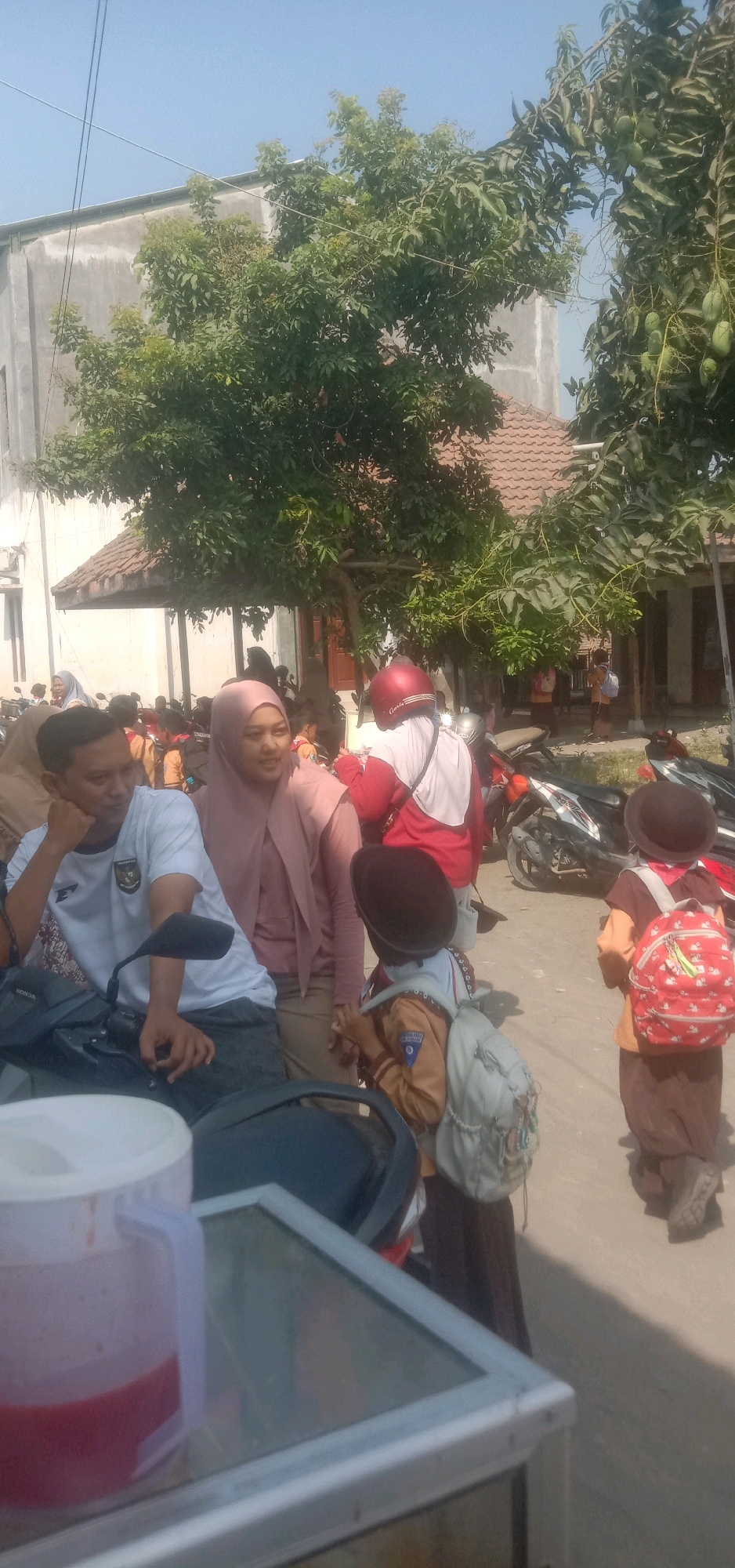 The image captures a sunny daytime scene on a street in Cangkringrandu, Indonesia, near a building with a tiled roof and some trees. In the foreground, a man in a white t-shirt is sitting on a motorcycle. To his right, a woman in a pink hijab is standing. Two young children wearing school uniforms and hats are also visible. One child, facing away from the camera, has a light-colored backpack, while another child in the background has a red backpack with a pattern. Several motorcycles are parked in the background. A large plastic pitcher containing a red liquid is in the lower left corner of the frame.