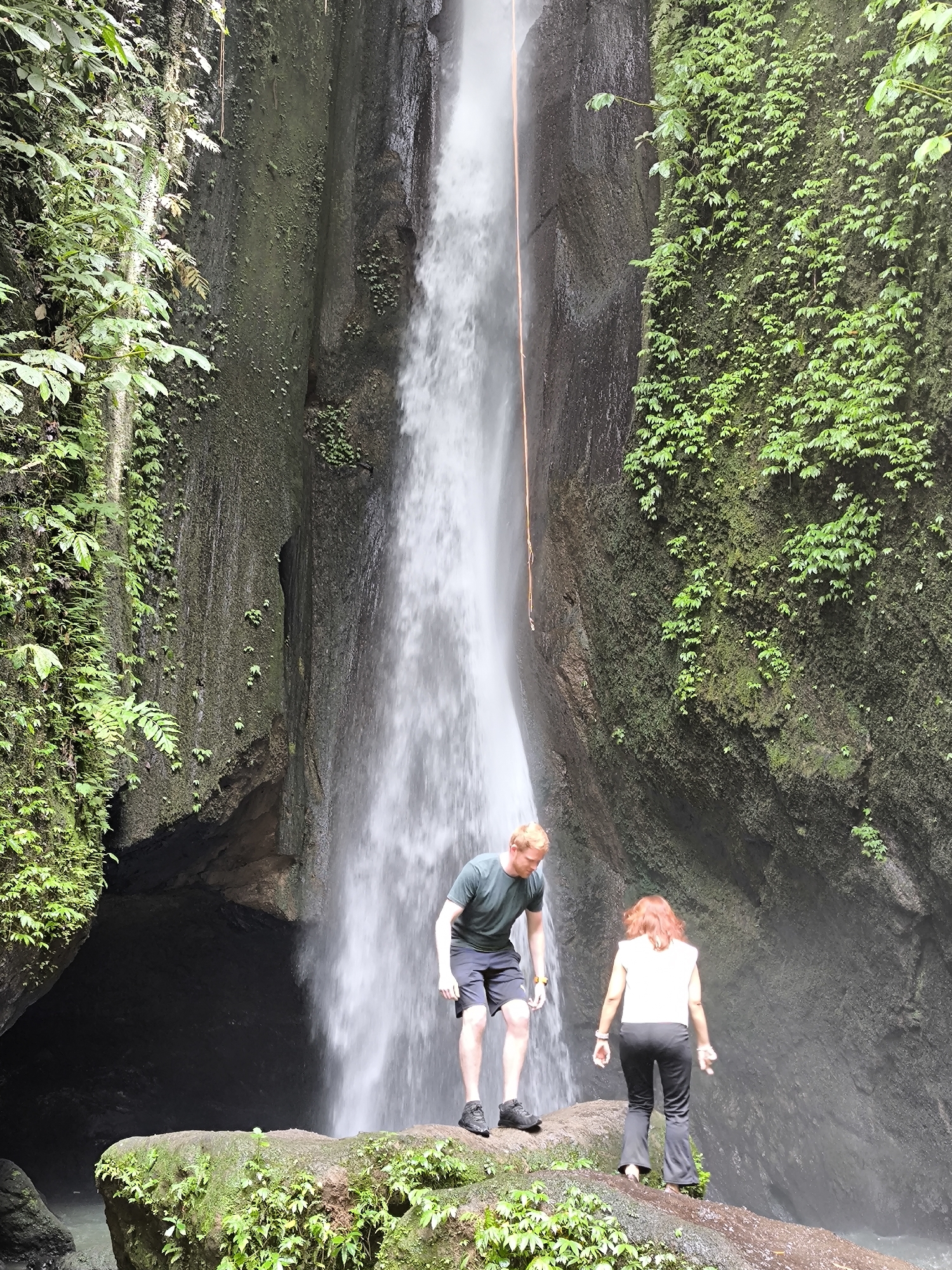 A medium shot depicts two individuals standing on a moss-covered rock formation at the base of a waterfall. The waterfall, located in Mekarsari, Indonesia, cascades from a significant height between two rock faces densely covered with green vegetation. An orange rope hangs down the center of the waterfall. The man on the left wears a green T-shirt and dark shorts, and has red hair. The woman on the right wears a white shirt and black pants. Both individuals are looking down. The environment consists of wet rock, moss, and lush greenery.