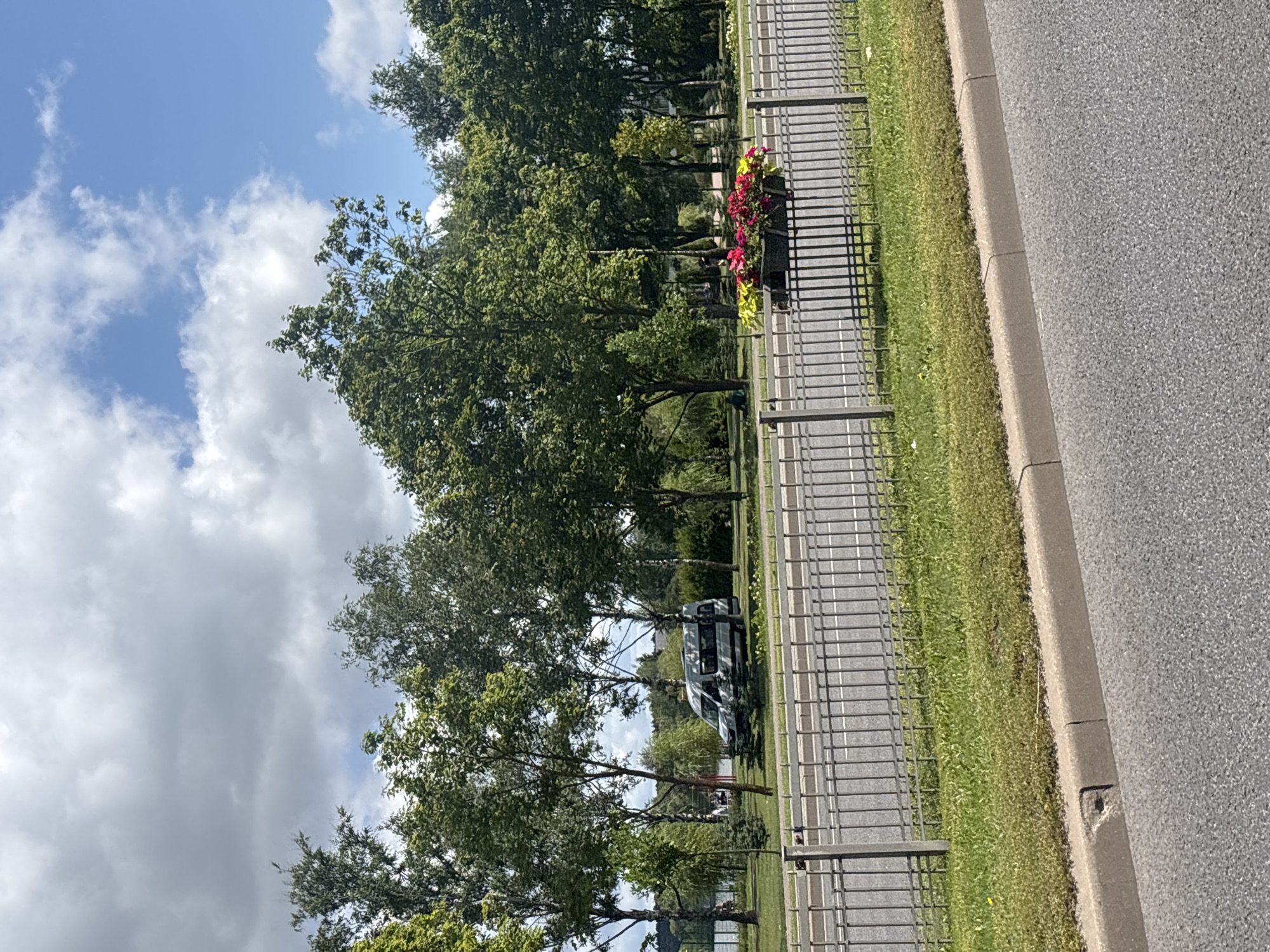 The image shows a scene in Minsk, Belarus, with a road in the foreground separated from a grassy area by a metal fence. A planter box with red and yellow flowers is visible behind the fence, and a grey van is parked among trees on the grassy area. The sky is partly cloudy with patches of blue. The road has visible lane markings. The environment appears to be a park-like setting with mature trees and well-maintained greenery.