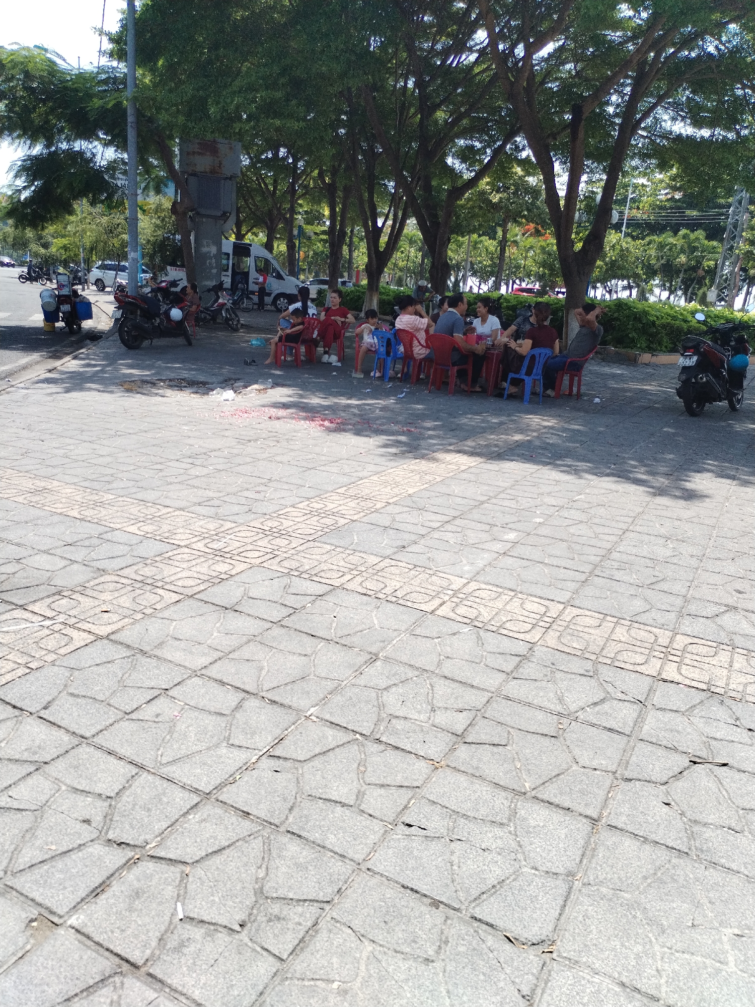 In the city of Bac Nha Trang Ward, Vietnam, a group of people are seated at a table on a paved sidewalk. The group consists of approximately seven individuals, a mix of adults and possibly children, some of whom are using red and blue plastic chairs. They appear to be engaged in a meal or a social gathering. In the background, several motorcycles are parked, and a white van is visible. The scene is set against a backdrop of lush green trees, suggesting an outdoor urban environment. The pavement is made of rectangular stone tiles with a decorative pattern. There is some red debris scattered on the ground near the table.