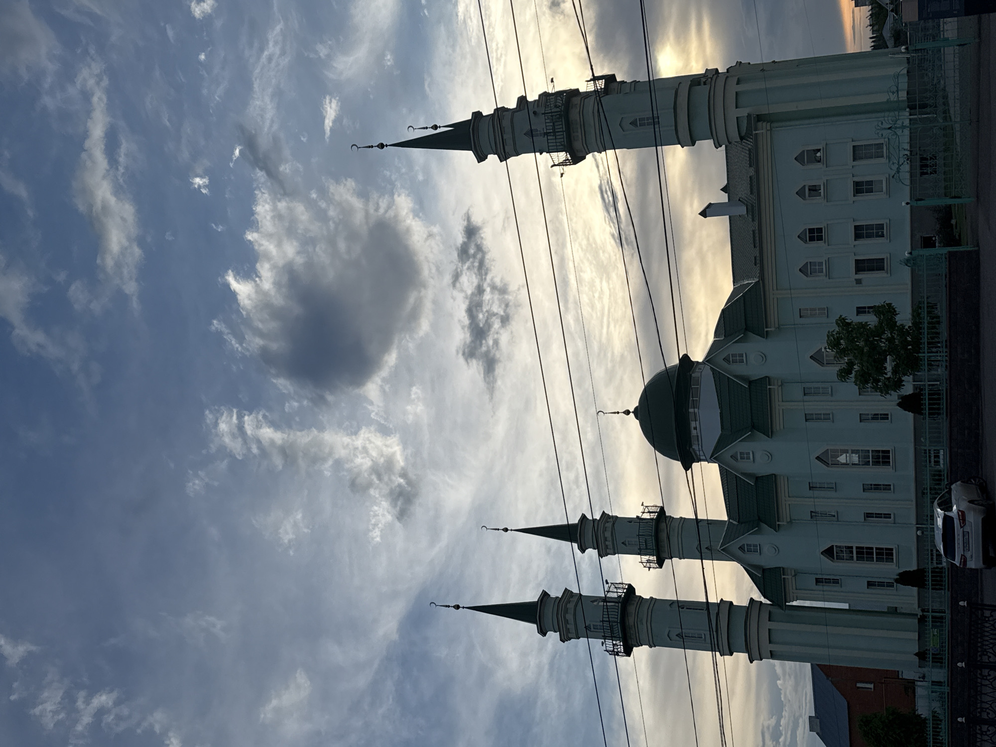 The image shows a mosque with its distinctive light blue facade and green domed roof, flanked by two tall, slender minarets. The mosque is a significant architectural feature in the urban landscape, its spires reaching towards a dynamic sky filled with a mix of dark and light clouds. Power lines are visible in the foreground, stretching horizontally across the frame, partially obscuring the upper portions of the minarets. A white car is parked in front of the mosque, its rear facing the camera, suggesting a public space. The building's facade is detailed with windows and decorative elements, typical of Islamic architecture. The overall impression is of a serene religious structure set against a dramatic sky, possibly at dusk or dawn given the lighting conditions. The scene is set in an urban environment, indicated by the presence of buildings and infrastructure like power lines and a fence.