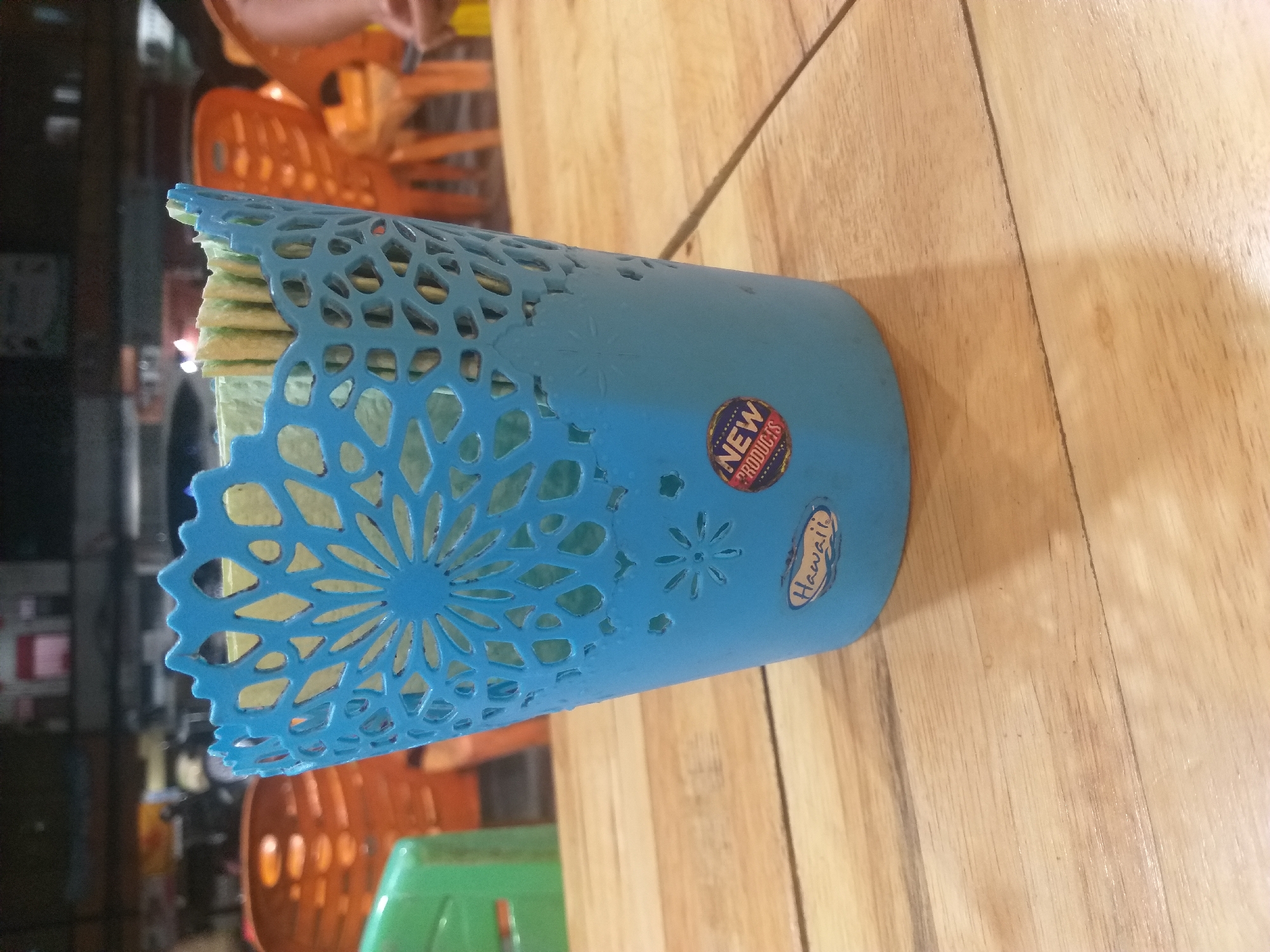 A close-up, eye-level, medium shot captures a light blue plastic basket with an ornate, perforated design, resembling a flower or snowflake pattern, on its upper rim. The basket is filled with what appear to be individually wrapped snacks or food items, visible through the perforations at the top. The basket is adorned with two stickers: a small oval sticker on the front with the word "Hawaii" and a round sticker below it featuring the text "NEW PRODUCTS" in a circular arrangement. The basket rests on a wooden table with a visible grain. In the background, out of focus, are several orange plastic chairs and a glimpse of a green plastic chair. The overall setting suggests an indoor environment, potentially a cafe or a casual eatery, possibly in Dumai City, Indonesia, given the context provided, though no explicit location markers are visible in the image itself. The lighting is moderate, casting a soft shadow from the basket onto the table.