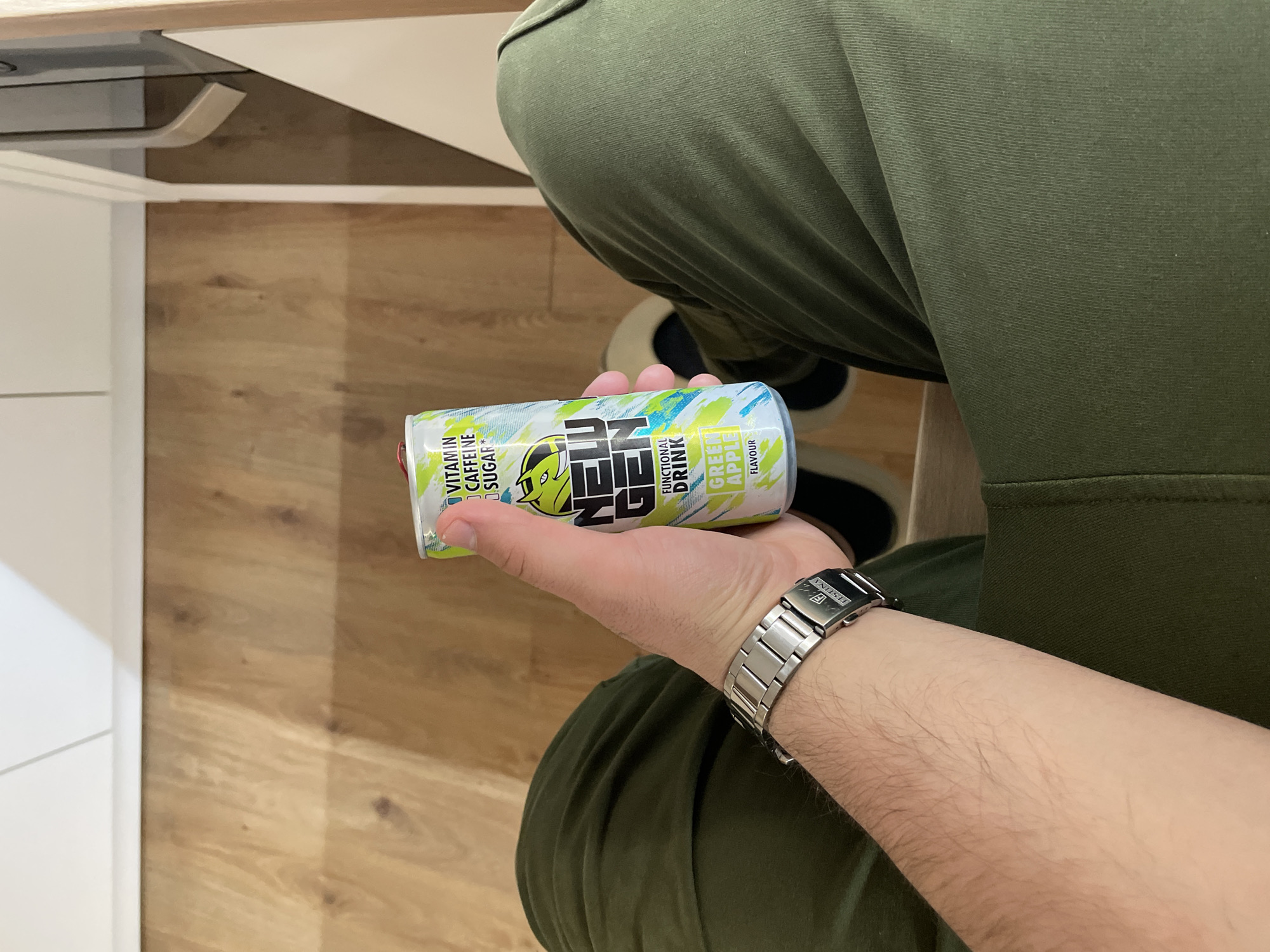 A first-person perspective shows a person wearing a silver wristwatch holding a green and blue energy drink can. The can features the word "NEU" in large black letters. The person is wearing olive green pants. The background consists of a light wood floor and a partially visible white table. The location is unknown.