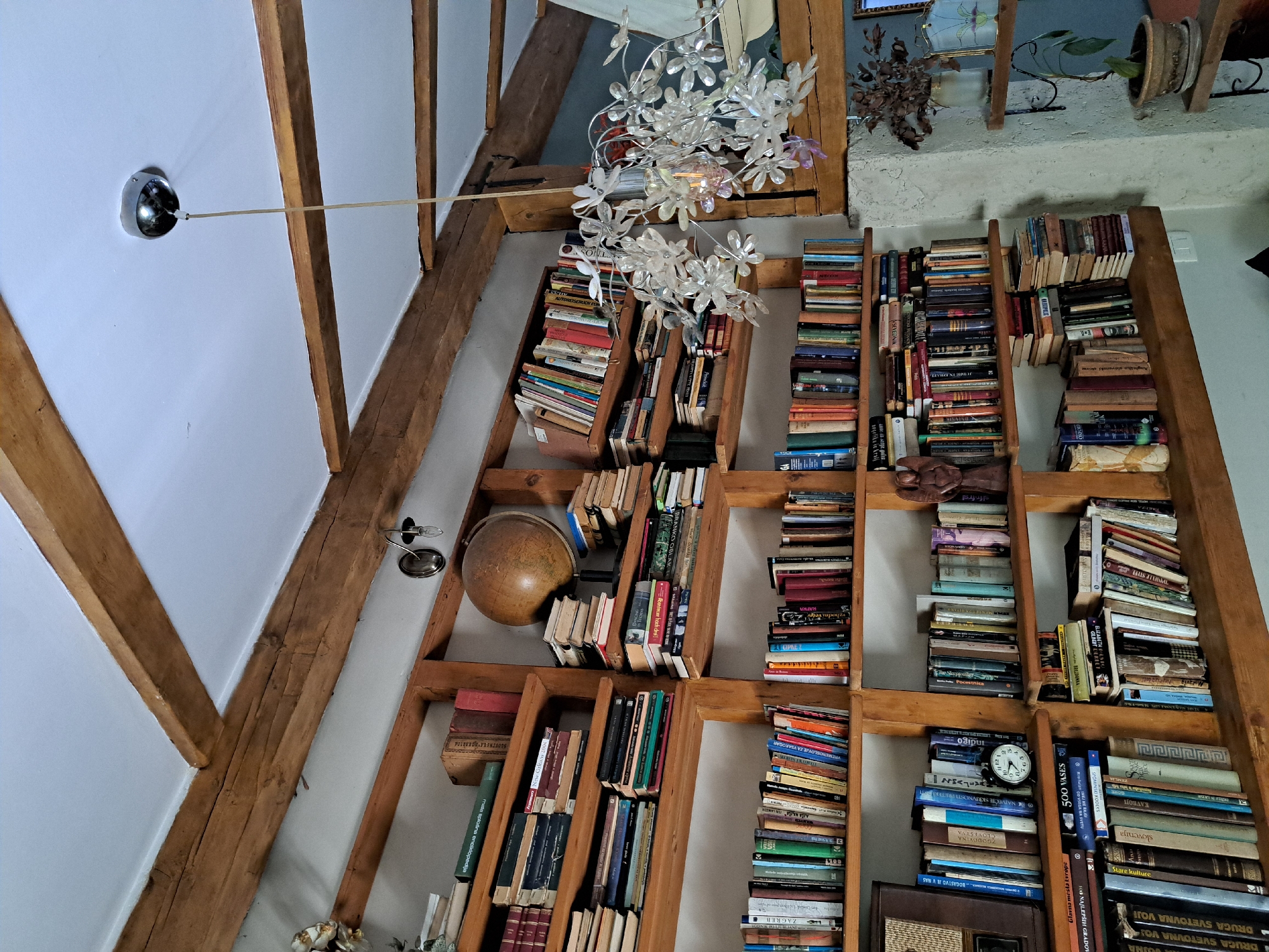 The image shows a room with wooden beams on the ceiling and white walls. There are several wooden bookshelves filled with books of various sizes and colors. On one shelf, a globe is visible. A chandelier with flower-shaped ornaments hangs from the ceiling. On a different shelf, a small analog clock is placed next to a collection of books. In the background, a statue of an angel is present. There are also plants in pots on shelves. The overall impression is of a cozy, well-stocked library or study. The scene appears to be indoors, possibly a home.