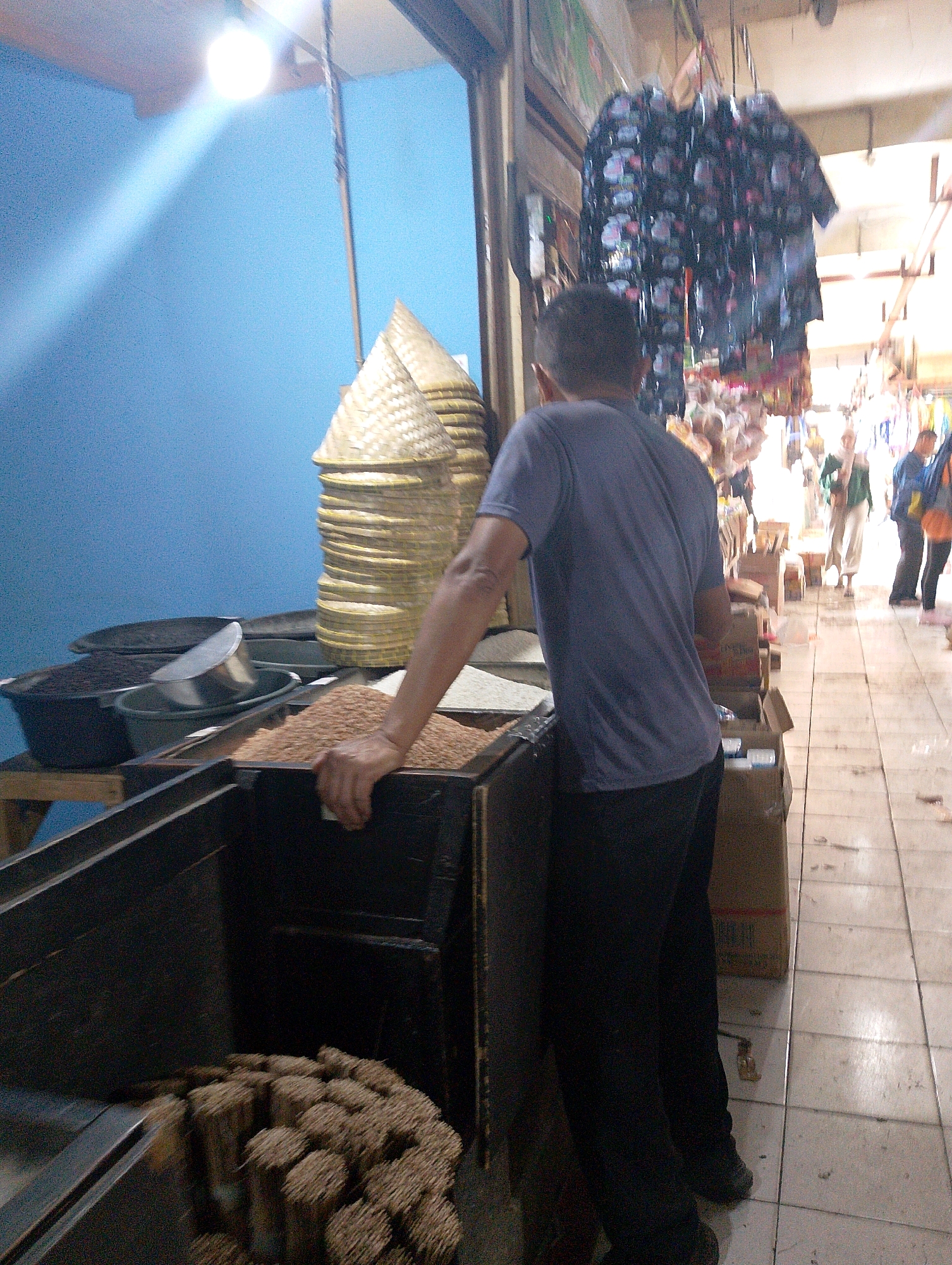 This is a full-shot, eye-level, slightly blurry photograph taken in a market stall in Lembang, Indonesia. The scene is brightly lit with a large bulb hanging from the ceiling casting a strong shaft of light to the left. In the foreground, a man wearing a grey t-shirt and black pants is standing with his back to the camera, his right hand resting on a wooden counter. The counter contains two dark, rectangular bins. The left bin appears to hold a pile of what looks like incense sticks, while the right bin contains some sort of grain or seed. Behind the man, a tall stack of woven conical hats is visible. The background shows other market stalls and shoppers, with bright lighting and some merchandise hanging from the ceiling, including what appear to be patterned bags or garments. The floor is tiled with a grid pattern. The overall impression is a busy, everyday market environment.