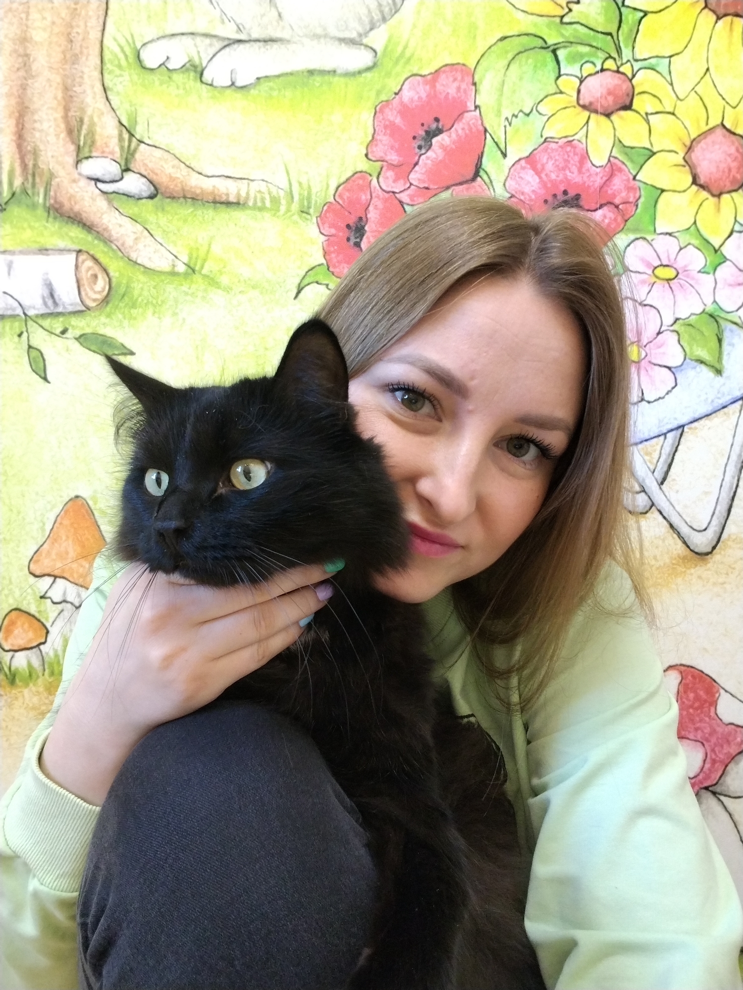 A woman with light brown hair is holding a black cat in her arms. The cat has green eyes. The woman is wearing a light green sweatshirt and dark pants. The background is a mural depicting flowers, trees, and mushrooms. The location is Chaikovsky, Russia.