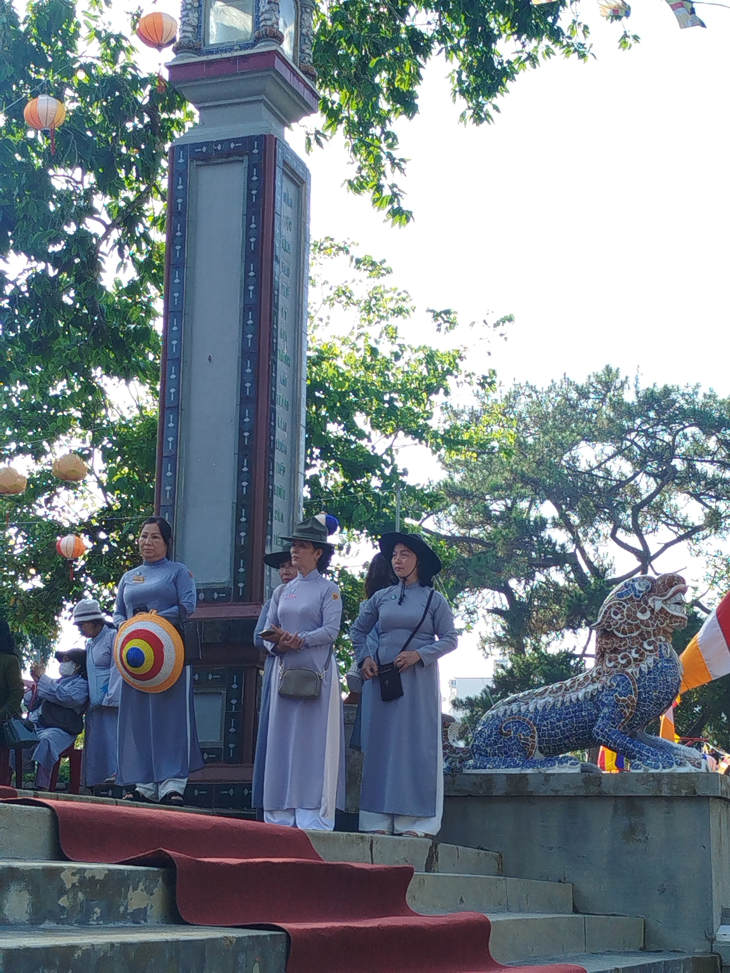 Captured from a low angle, the image depicts a scene set in Da Lat, Vietnam, featuring four women dressed in traditional Vietnamese garb (áo dài) with white pants and lavender tops. They stand on a tiered platform adorned with a red carpet, positioned behind a tall stone pillar decorated with symbolic motifs and flanked by a mosaic lion statue. The pillar is surrounded by trees, and hanging paper lanterns are visible within the branches. Other people are in the background.