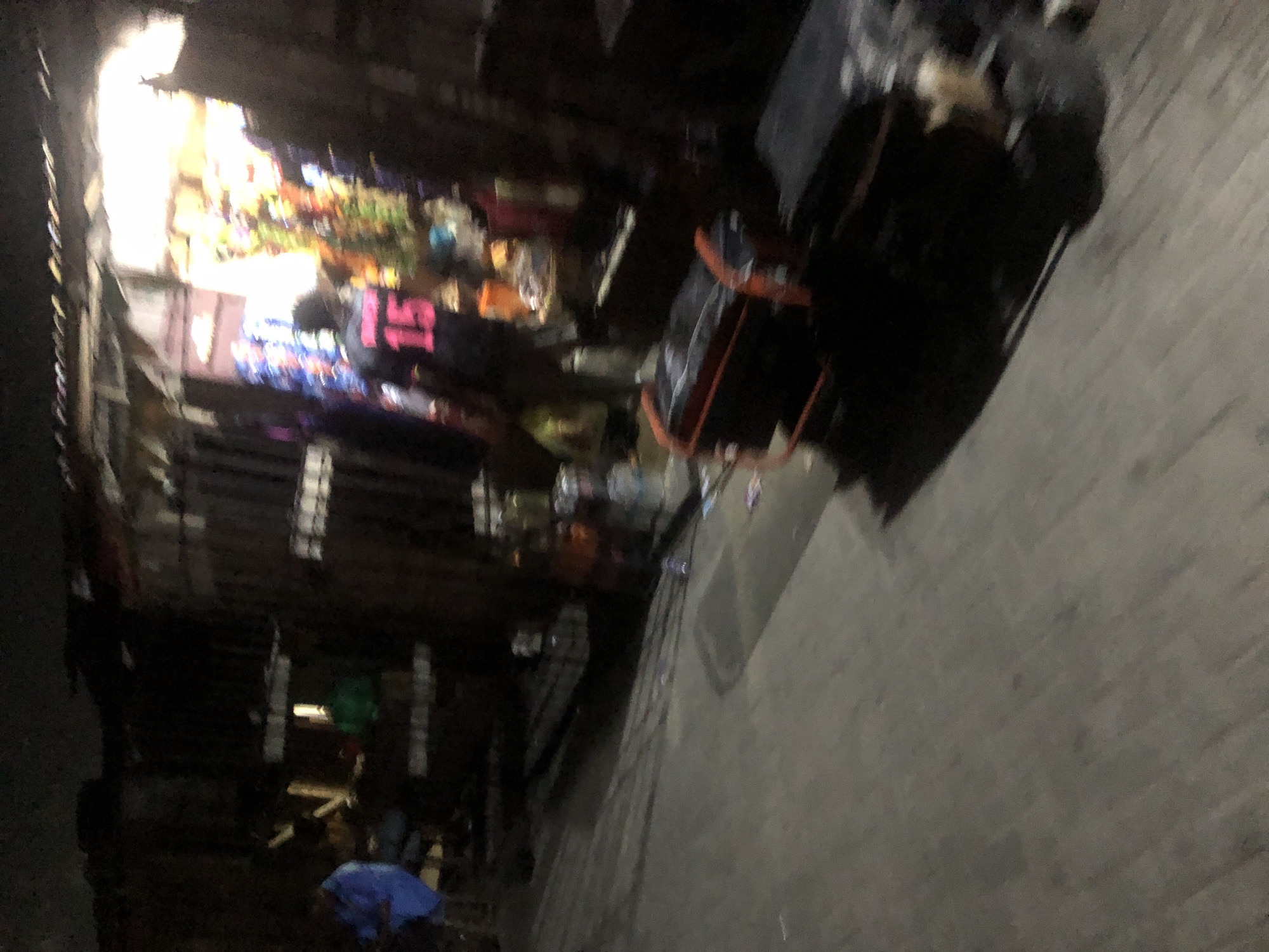 The image depicts a partially obscured street scene, possibly in Ado, Nigeria, given the prompt. A person wearing a shirt with the number "15" on the back is visible near a brightly lit shop. The shop appears to have a variety of goods on display. To the right, there is a black object, which looks like some type of luggage on wheels, situated on what seems to be a concrete walkway or road. Another figure is lying on the ground further down the street and is covered by a blue sheet or covering. The overall lighting is dim, and the image is out of focus.