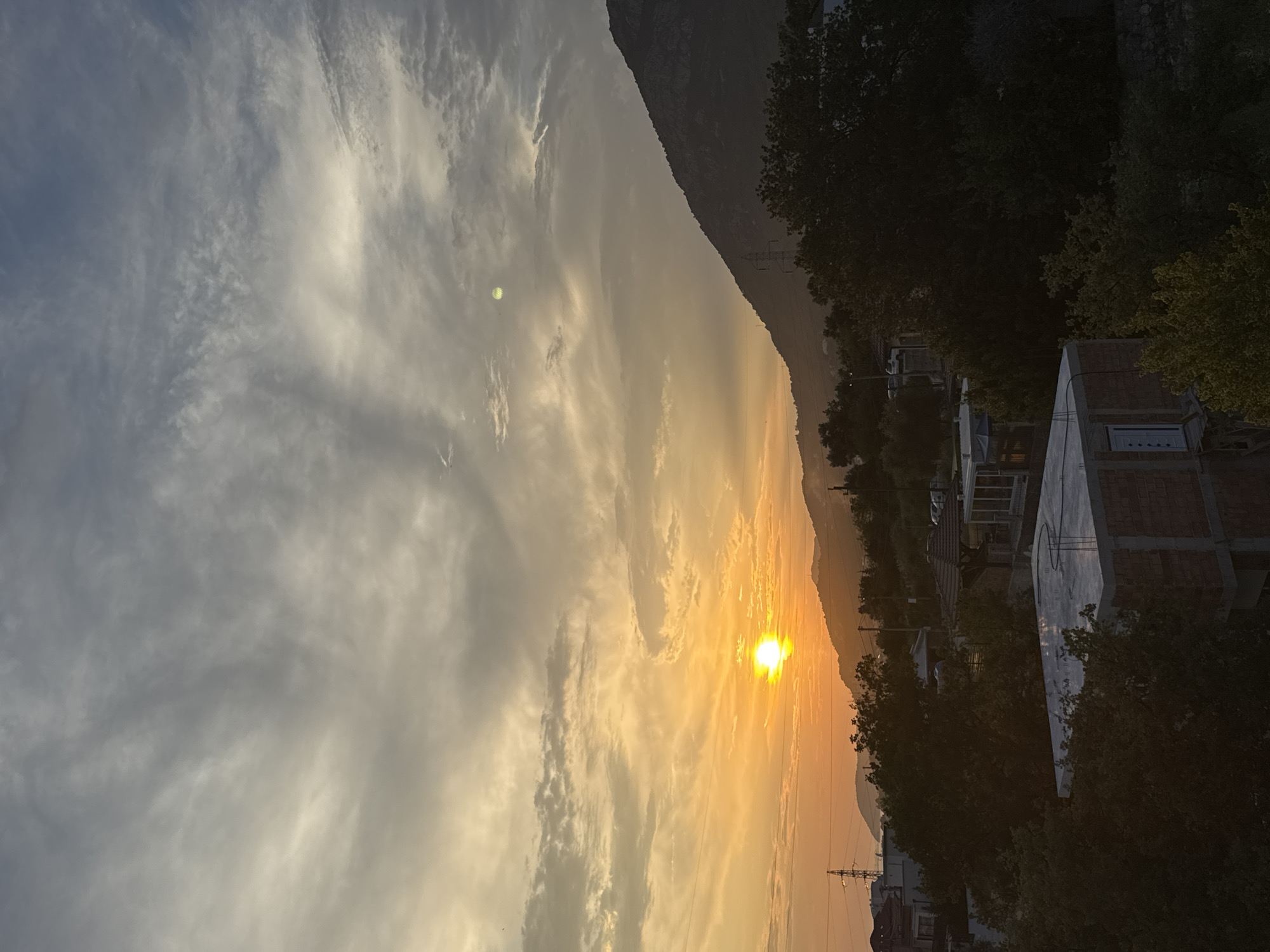 The image depicts a sunset over a hillside landscape in Sustaš, Montenegro. The sun, a bright orb, is positioned on the left side of the frame, casting a warm orange glow across the sky and illuminating the clouds with golden hues. The sky is a mix of oranges, yellows, and grays, with wispy clouds stretching across the upper portion of the image. A dark, silhouetted mountain range dominates the right side of the frame, with its peak rising into the fading light. In the foreground, the landscape features scattered buildings, some under construction, interspersed with dense foliage. Electrical pylons are visible in the distance, suggesting the presence of infrastructure within the mountainous terrain. The overall scene captures a serene, end-of-day atmosphere.