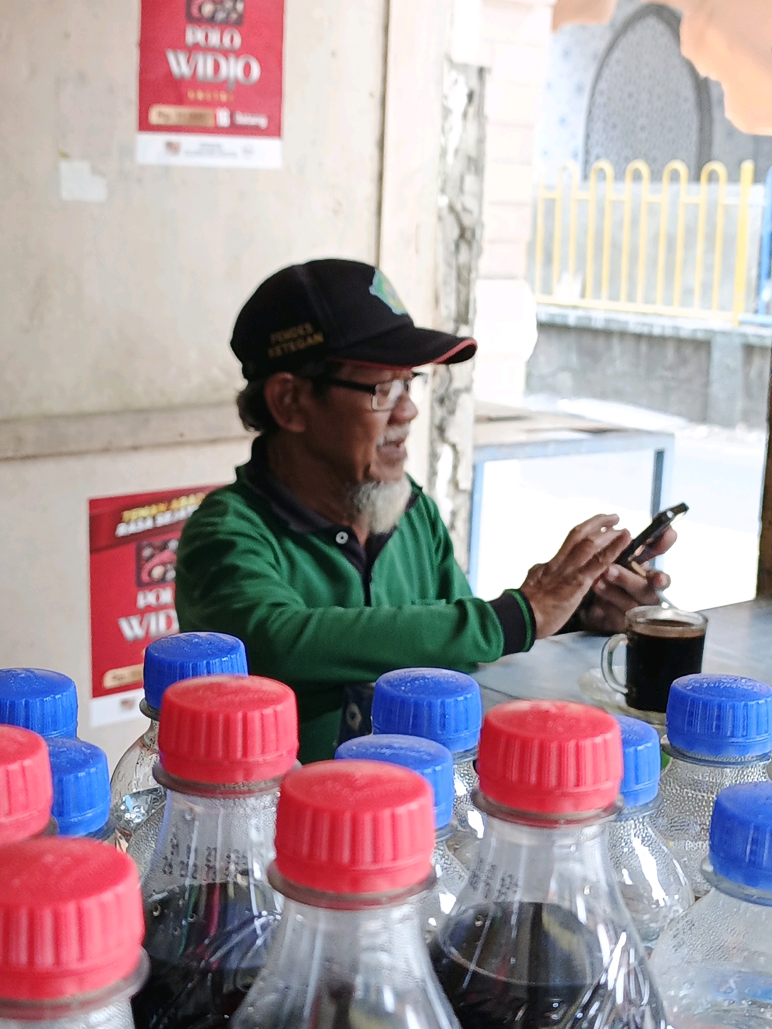 In Ketegan, Indonesia, a man wearing a black baseball cap and glasses sits at a table, looking at his smartphone. He is wearing a green long-sleeved shirt. In front of him on the table is a clear mug filled with a dark liquid, presumably coffee, and a white saucer. In the foreground, several plastic bottles with red and blue caps are visible. Behind the man, a red sign with white lettering, "POLO WIDIO", is partially visible on a wall. The environment appears to be an outdoor or semi-outdoor setting with a yellow metal fence in the background.