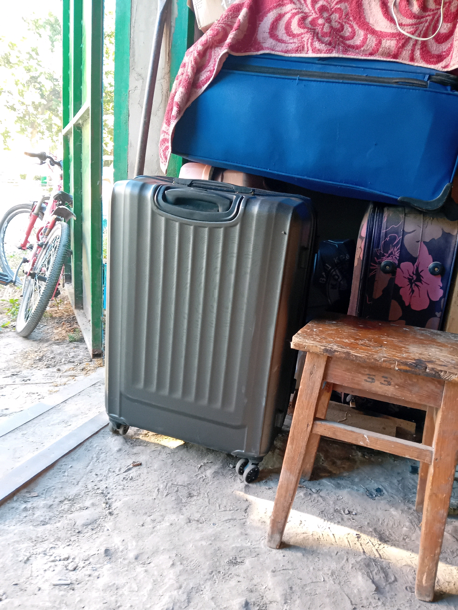 This image depicts an outdoor storage area with several pieces of luggage and a bicycle. In the foreground, a large grey suitcase with ridged detailing and black wheels stands upright. To its right, a small wooden stool with worn surfaces and the number "55" stenciled on it is positioned. Behind the stool and to the right of the grey suitcase, another suitcase with a floral pattern of pink and purple flowers on a dark background is visible. Further back, a large blue suitcase is stacked on top of other items, and a red and white patterned towel is draped over it. To the left of the scene, a red bicycle with black tires is leaning against a green metal gate. The ground is covered in dirt and debris. The scene appears to be located in an outdoor storage area in the city of Chuhuiv, Ukraine, possibly near a residential building or a communal storage facility.