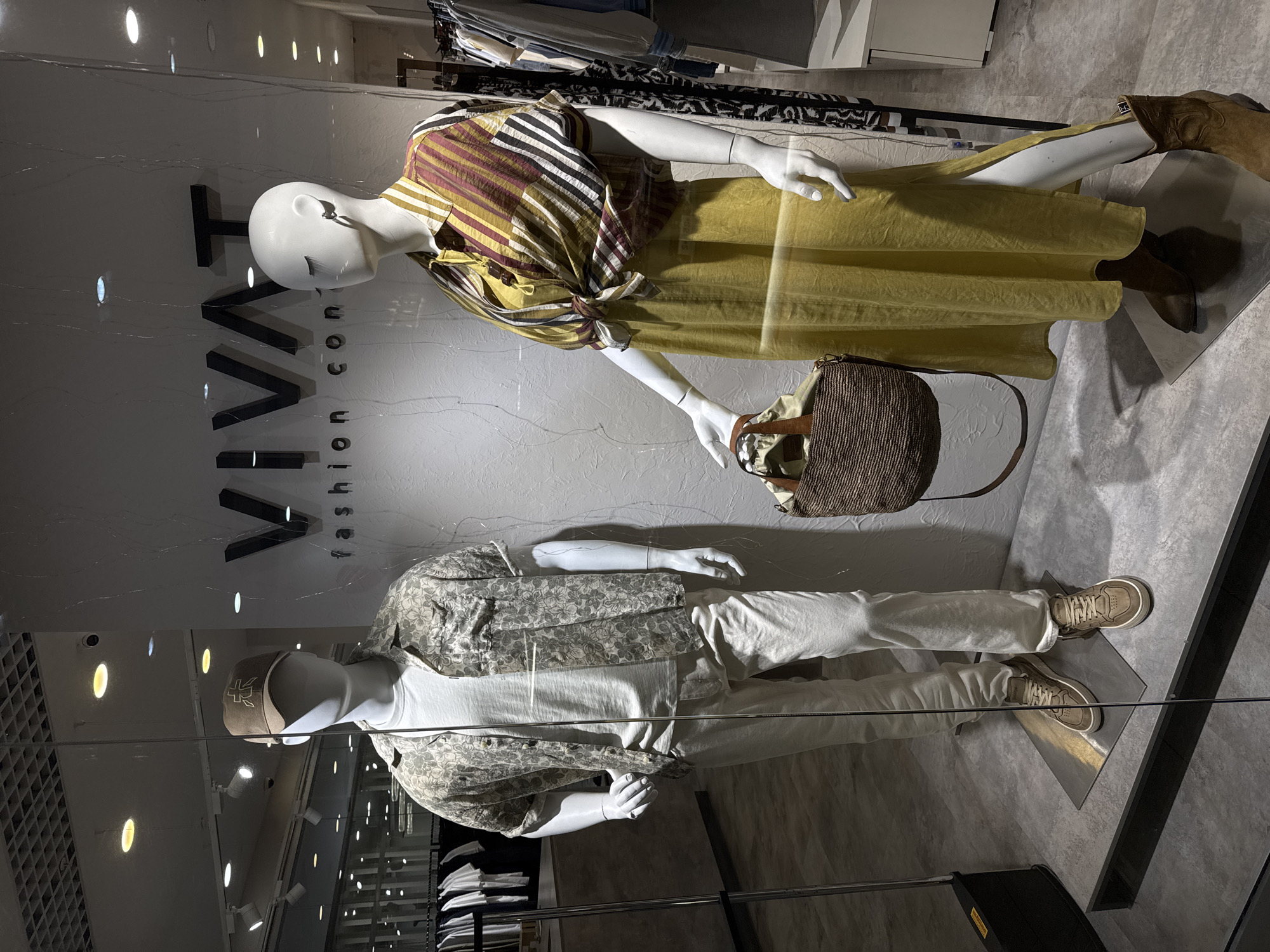 A store window display, located in an unknown city, features two mannequins. One mannequin is dressed in a horizontally striped, long-sleeved shirt, tied at the waist, paired with a light yellow dress. This mannequin also wears brown boots and carries a brown woven bag. The second mannequin wears a short-sleeved button-down shirt with a floral pattern over a white t-shirt, white pants, and tan sneakers. The store's name, "IVAVI fashion concept," is visible on the glass of the window. Other clothing items are visible on racks in the background.