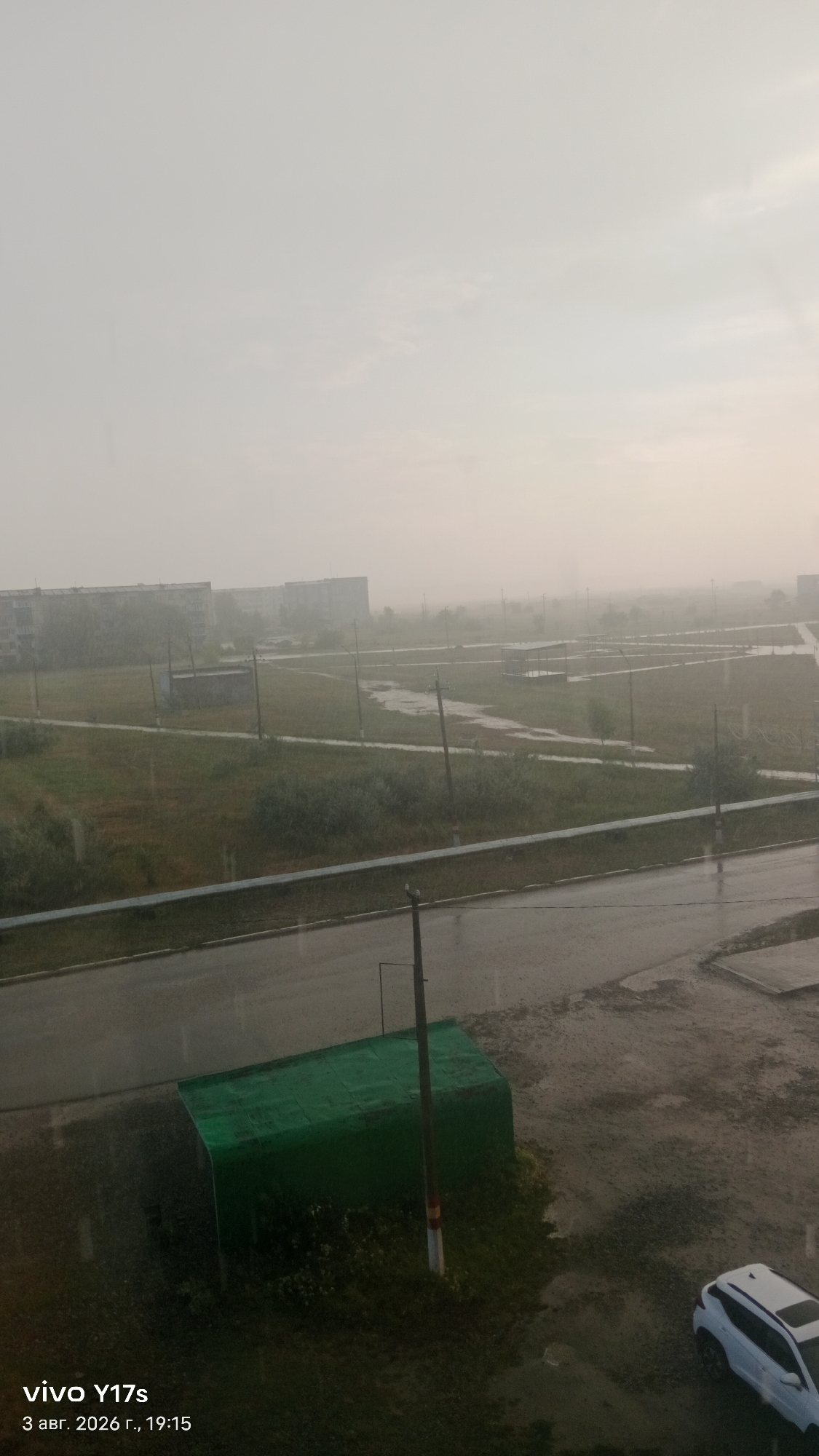 This is a medium-angle, eye-level shot of a rainy day in the city of Rudny, Kazakhstan. The foreground shows a wet, paved area with a small, green structure resembling a shed. A light pole stands next to the shed. In the middle ground, a road runs horizontally, bordered by a railing. Beyond the road is a grassy area with scattered trees and pathways, and in the background, several apartment buildings are visible through the heavy rain and mist. The sky is overcast and hazy. A white SUV is parked on the right side of the foreground. The timestamp in the bottom left corner reads "vivo Y17s 3 авг. 2026 г., 19:15".