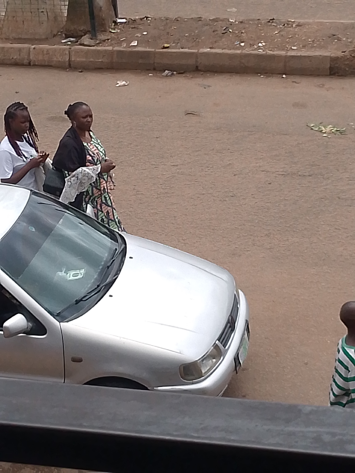 The image captures a street scene in Bukuru, Nigeria. Two women are walking on the dirt road near a silver Volkswagen Polo. The woman on the left has her hair in braids and is wearing a white t-shirt, holding a mobile phone. The woman on the right has her hair tied back and is wearing a black jacket over a colorful, patterned dress with white lace trim on the sleeves. A young child in a green and white striped shirt is visible on the far right of the image, facing away from the camera. The road is unpaved and dusty, with some debris and scattered leaves. A concrete curb runs along the side of the road. The scene appears to be in an urban or semi-urban environment.