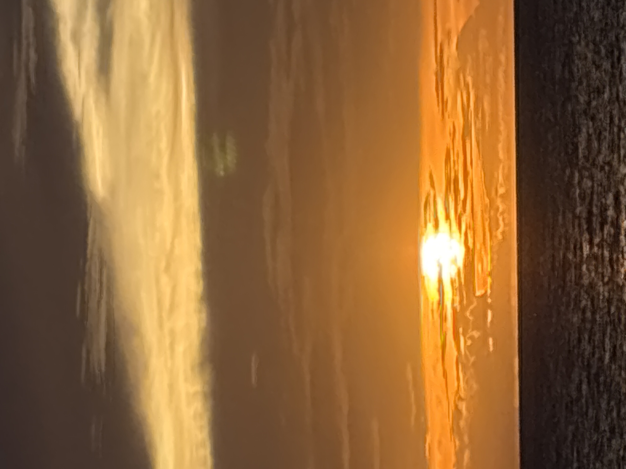 This media file shows a close-up, vertical shot of a translucent, textured surface with light shining through it. The surface appears to be made of some kind of glass or plastic, with vertical lines and undulations creating a rippled effect. To the left, the light is a soft, pale yellow, suggesting it's passing through a diffused material. Towards the right, the light intensifies to a bright, warm orange, with the texture of the surface becoming more pronounced. At the very edge on the right, there is a dark, rough, textured surface, possibly fabric or a rough wall, which appears to be in shadow. The overall impression is one of abstract light and texture. The lighting suggests it could be sunrise or sunset, with the warm orange glow being particularly strong. The orientation of the image is vertical, and the composition focuses entirely on the interplay of light and texture. There is no discernible text, people, or identifiable objects in the frame. The scene is abstract and evokes a sense of warmth and perhaps a gentle, ambient light source.