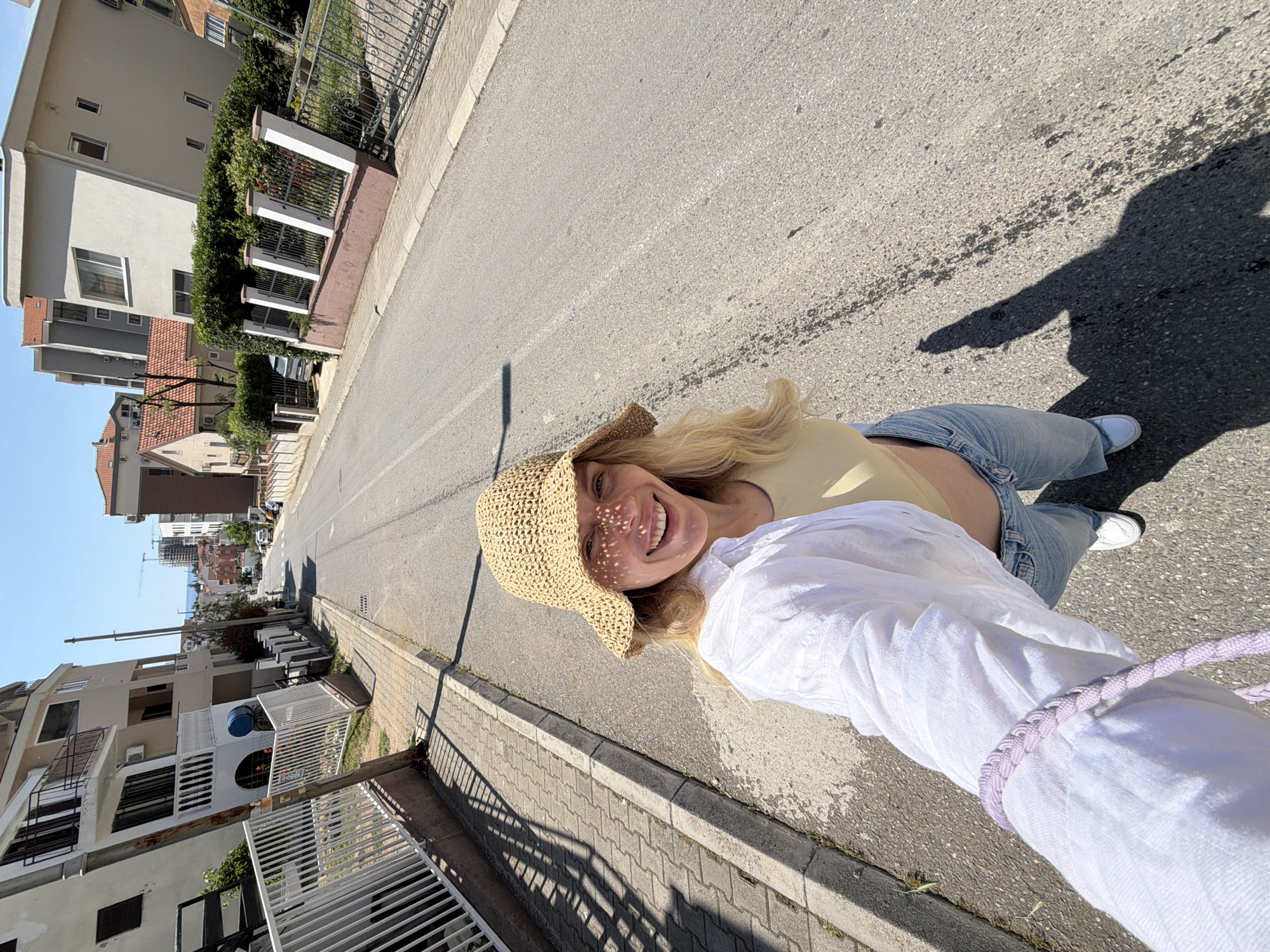 A cheerful blonde woman takes a selfie on a sunny day in Budva, Montenegro. She is wearing a straw hat that casts a dappled shadow on her face, a light yellow crop top, light blue jeans, and white sneakers. Her white long-sleeved shirt and a braided purple cord on her wrist are visible in the foreground. The woman is smiling widely, indicating a happy and relaxed mood.
The scene depicts a paved street with a concrete sidewalk and curb on one side, flanked by residential or commercial buildings. These multi-story structures feature light-colored facades, some with balconies and red-tiled roofs, characteristic of Mediterranean architecture. Lush green hedges and various fences, including white picket and ornate metal designs, line the properties. In the background, more buildings and visible construction cranes suggest an area undergoing development. The bright daylight and strong, defined shadows on the road indicate it is likely midday or early afternoon under clear skies. No discernible text is visible in the image.