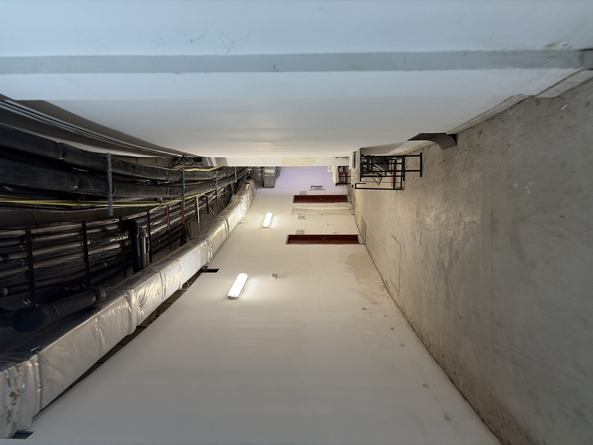The image depicts a view looking upwards in an interior space, likely a building's service shaft or stairwell. The perspective is tilted, giving a sense of looking up towards a distant ceiling.
On the left side of the frame, a dense array of black pipes and conduits runs along a wall. These pipes are insulated with silver-colored material in the foreground. Above them, a white ceiling extends across the top of the image.
The right side of the frame features a rough, concrete-like wall that extends upwards. Further up, against this wall, is a black metal structure, possibly a shelving unit or part of a ventilation system.
Along the center of the space, the white walls ascend. Several fluorescent light fixtures are visible, some illuminated. In the upper portion of the frame, there are doorways or openings set into the walls, some with dark red frames or doors. Faint water stains or discoloration are visible on the white surfaces.
The overall impression is one of functional, perhaps utilitarian, architecture. The lighting suggests it could be daytime, though the artificial lights are prominent. There is no visible text in the image. The scene is devoid of people, focusing solely on the architectural elements and infrastructure. The location context provided is Obzor, Bulgaria.