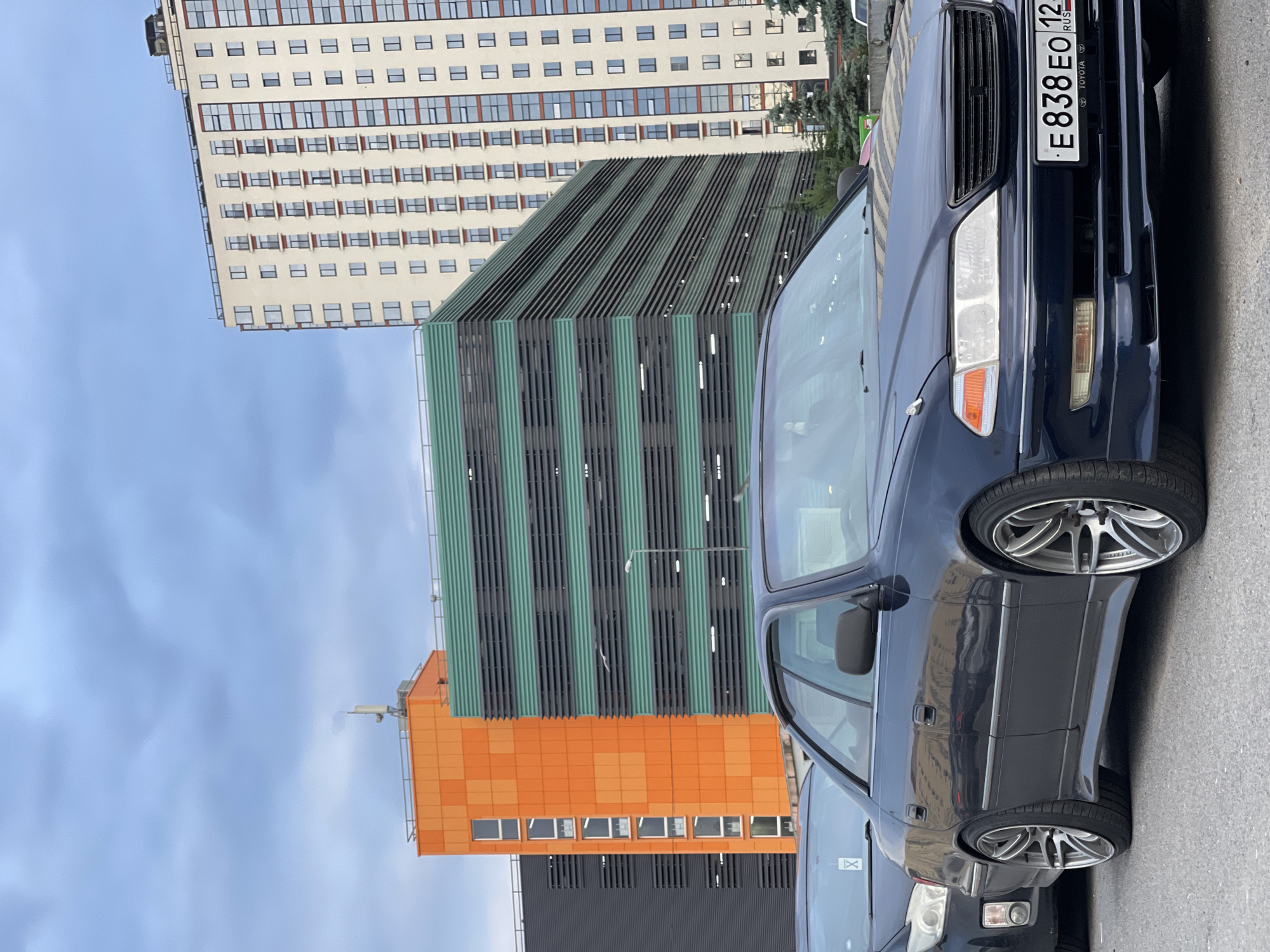 In an outdoor urban setting, a dark blue sedan is parked in the foreground, its front end facing the viewer. The car has chrome rims and a license plate that reads "E 838 EO 12 RUS". To the left of the sedan, the front of another dark-colored vehicle is partially visible. In the background, several modern buildings are prominent. A tall, cream-colored building with multiple windows and balconies stands on the right. In the center, a multi-story structure with horizontal green and black panels is visible, and to its left, an orange building with a similar panel design. The sky above is cloudy and overcast. Trees are present in the midground between the cars and the buildings. The ground appears to be asphalt.