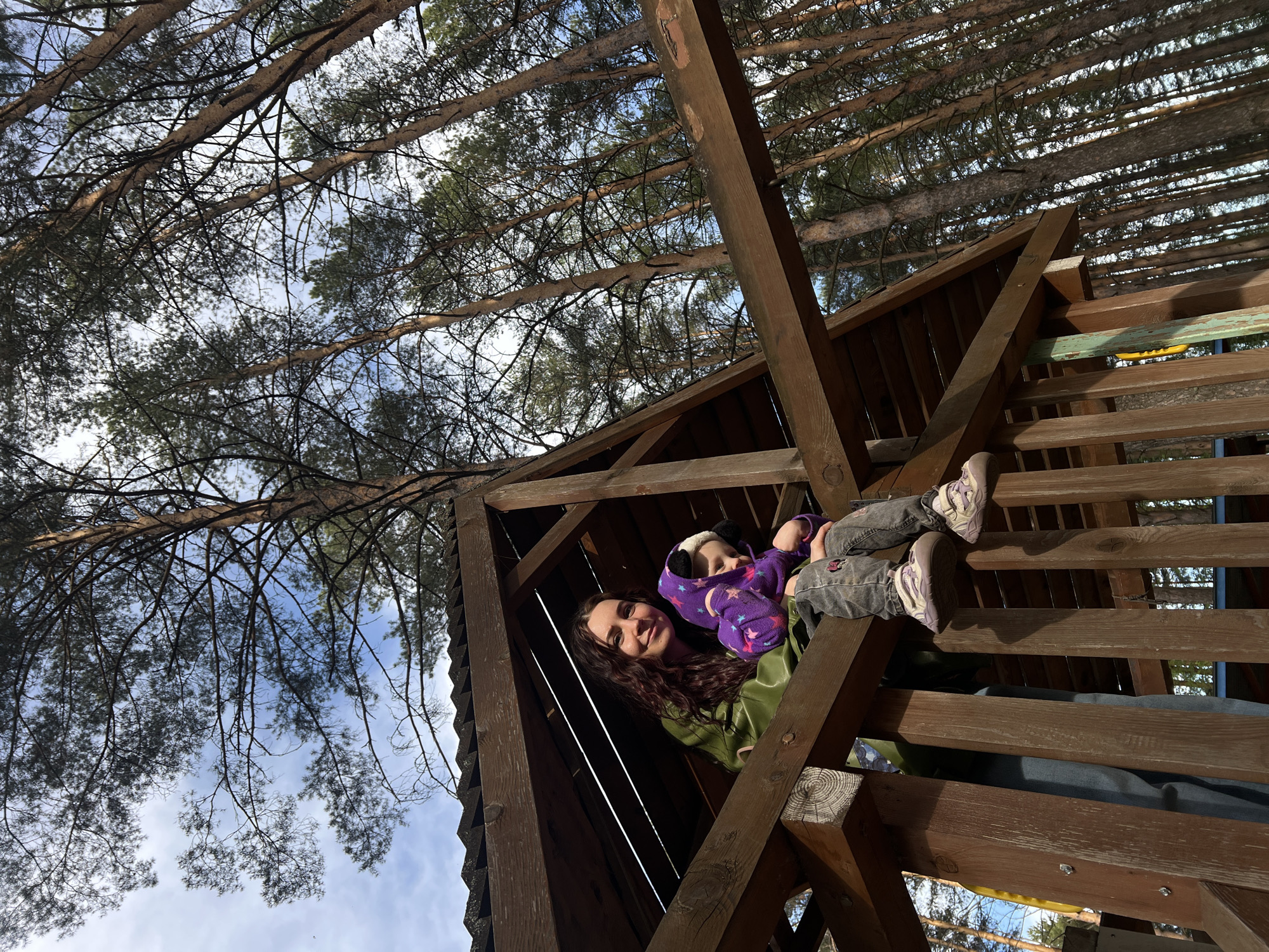 A low-angle shot captures a woman and child inside a wooden structure, possibly a playground or treehouse. The woman, with long, curly brown hair, wears a green jacket and smiles toward the camera. The child, wearing a purple hooded jacket with star patterns and panda ears, is partially obscured by the woman. The child is lying down with their feet propped up on a wooden railing, shod in lavender sneakers. The wooden structure is made of dark brown wood. The backdrop is a dense forest of tall trees with visible needles, as well as the sky with sparse clouds. The location is Chaikovsky, Russia.