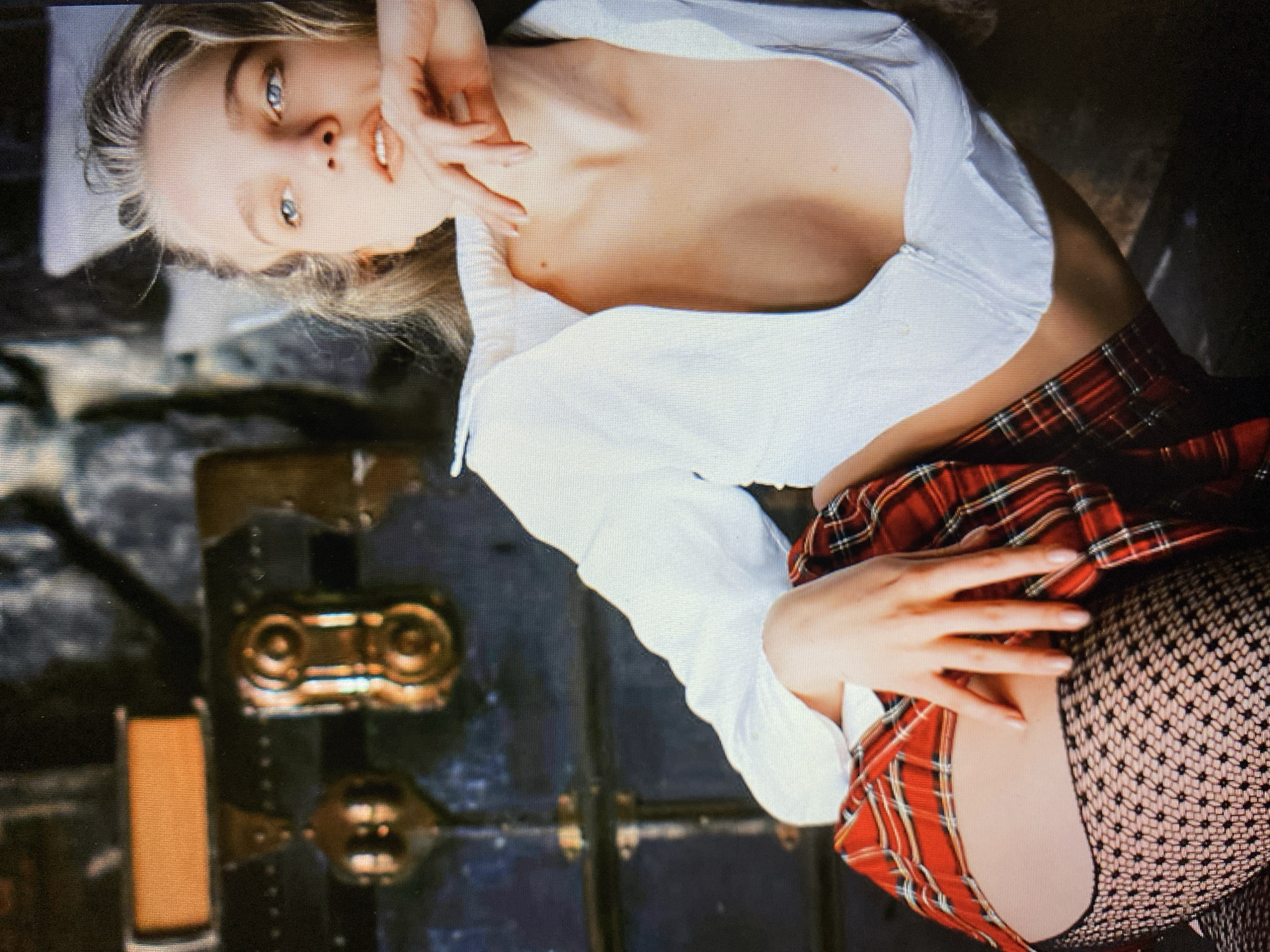 A young woman with blonde hair and blue eyes is lying on her side. She is wearing a white collared shirt that is unbuttoned, revealing her chest and a portion of her neck. Her right hand is resting on her face, and her left hand is near her hip, under her skirt. She is wearing a red plaid skirt and black fishnet stockings. The background is dark and appears to be a stone wall with some gold-colored decorative elements and a large, dark, textured surface. The image is a close-up, focusing on the woman's upper body and face. There is no information to determine the location in the city of Unknown, Unknown.