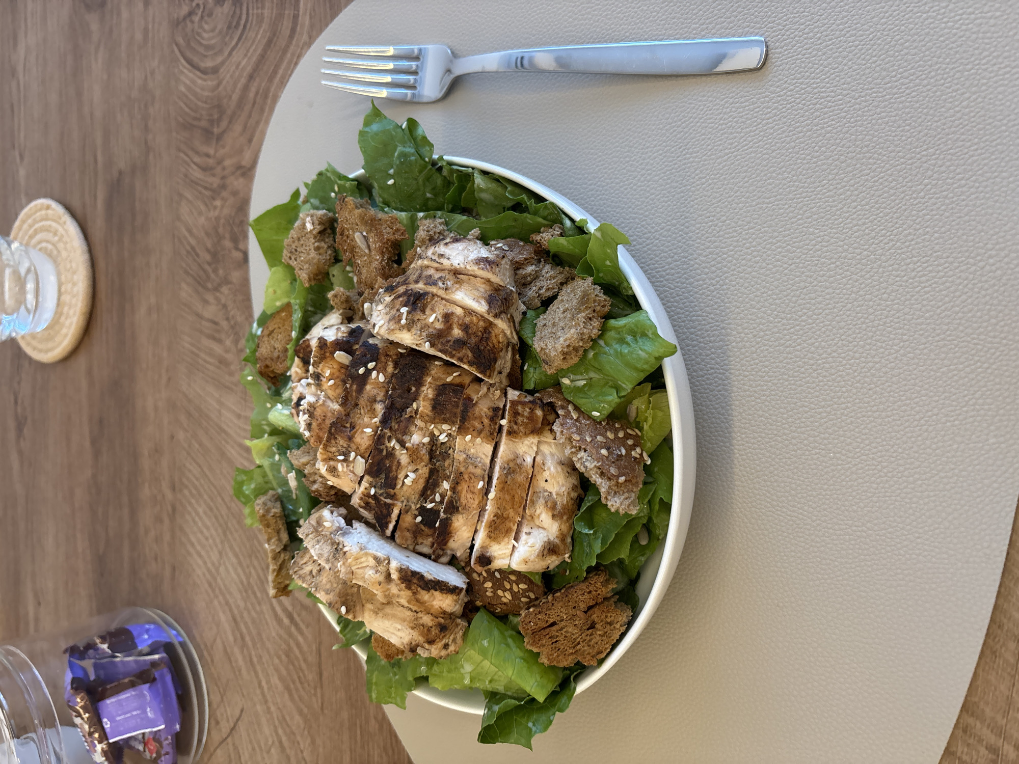 A close-up, high-angle shot shows a salad with grilled chicken and croutons in a white bowl. The salad is on a textured, light gray placemat, which is on a wooden table. A silver fork with a patterned handle rests to the right of the bowl. In the background, a glass jar filled with purple-wrapped candies and a coaster with a glass of water are visible. The scene is likely from a home dining area in Viernheim, Germany.